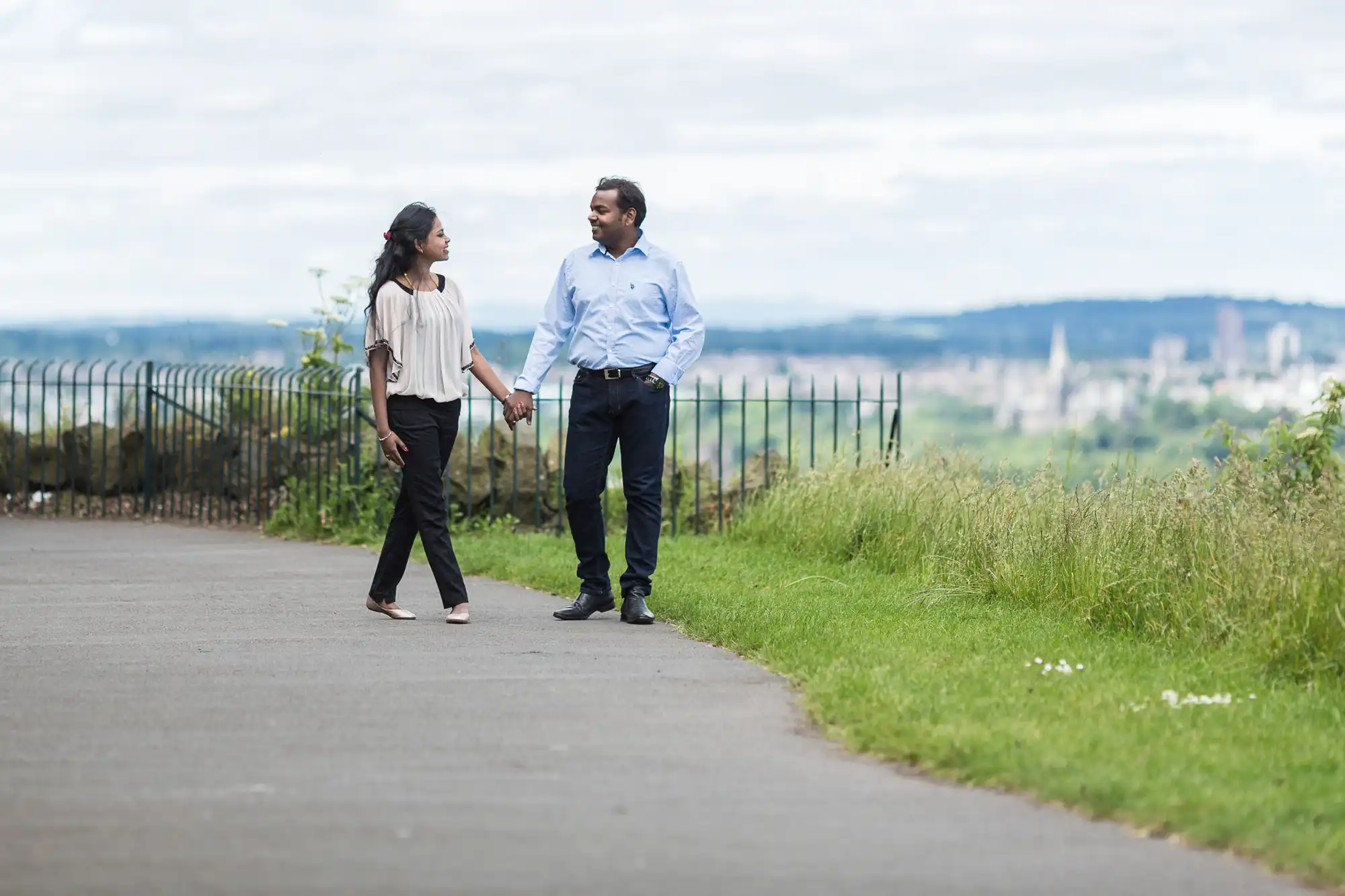 A couple holding hands while walking on a paved path, with a cityscape and greenery in the background.