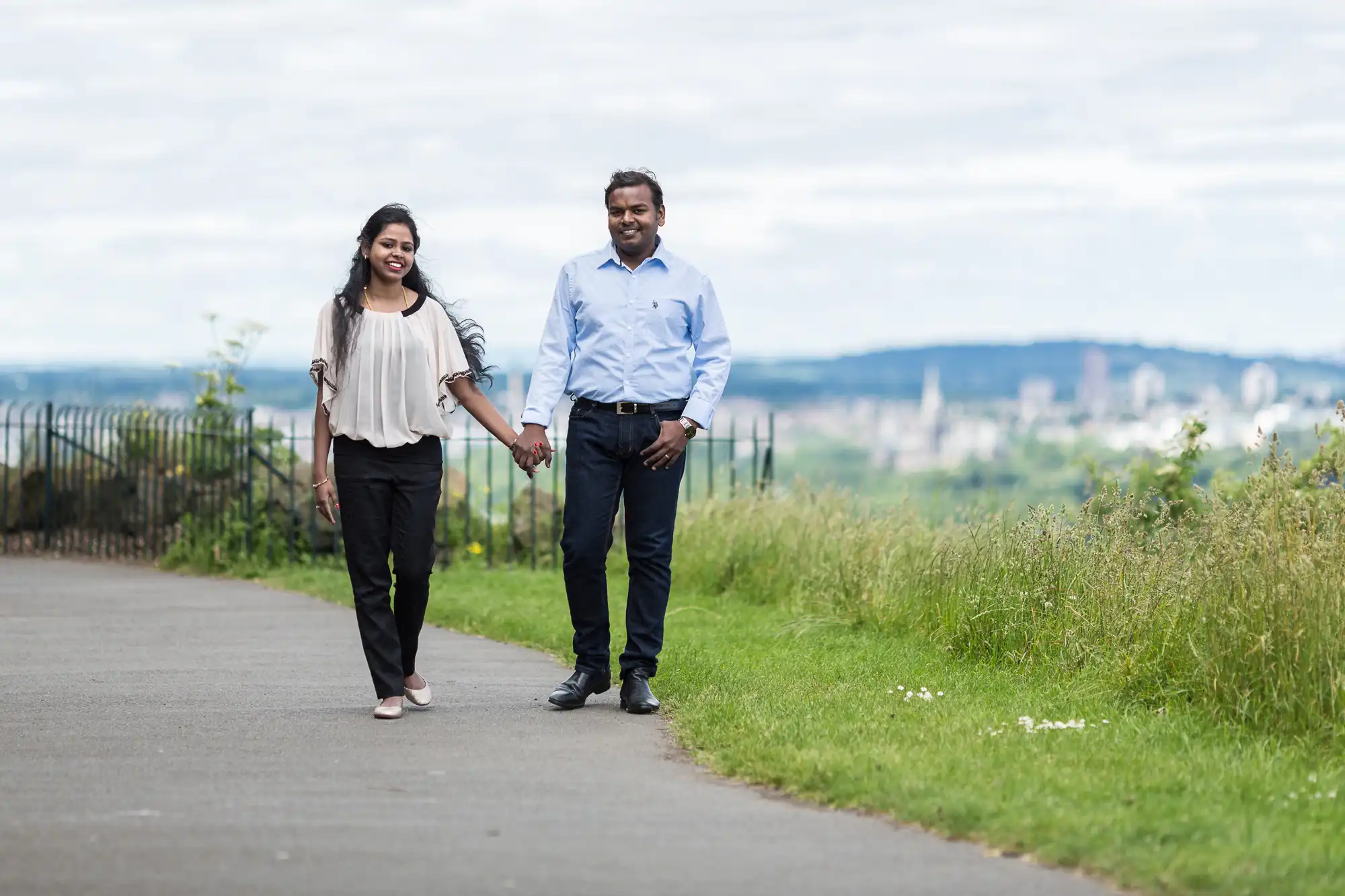 A man and woman hold hands while walking on a paved path with greenery and a cityscape in the background.