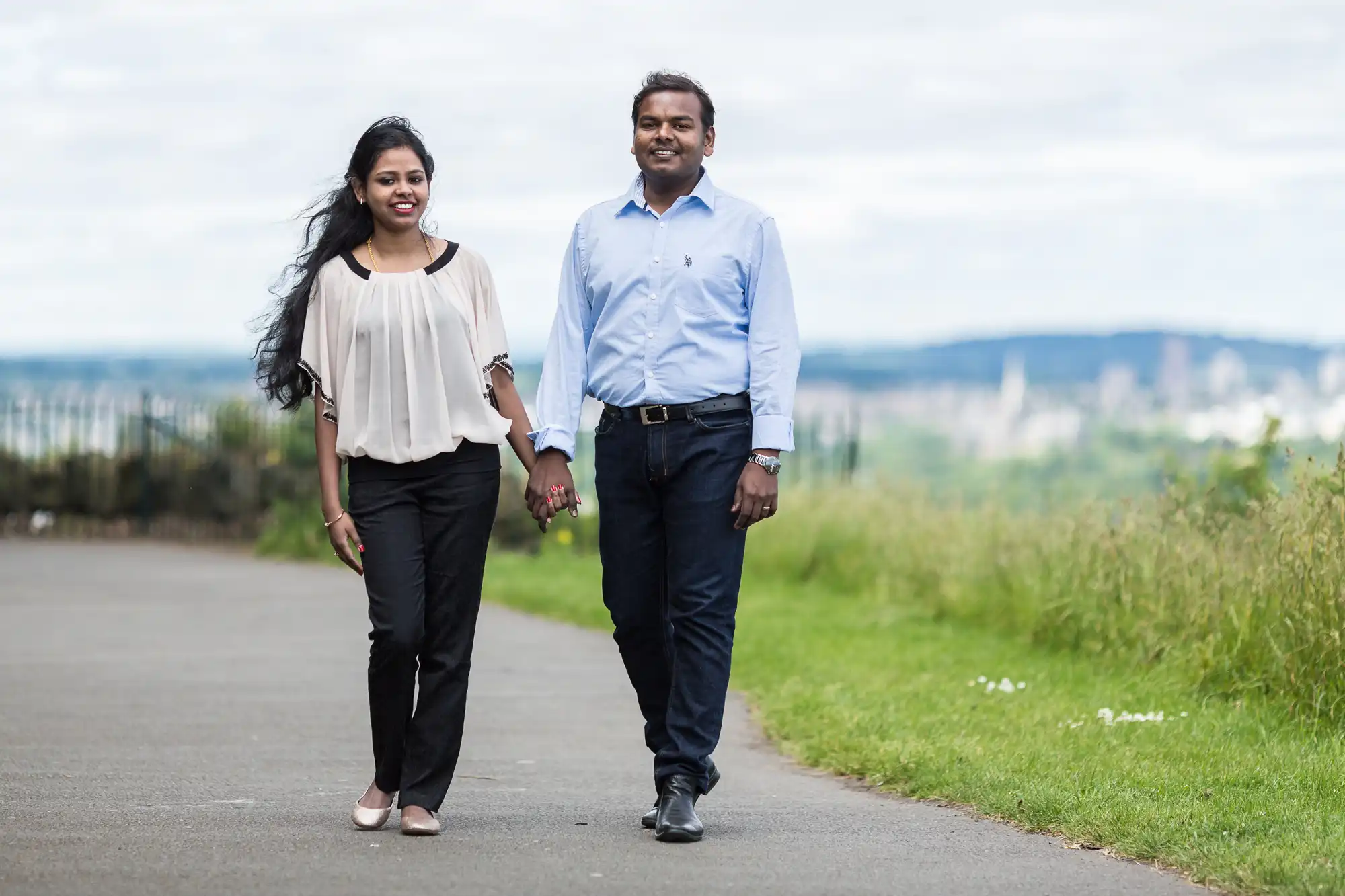 A couple holding hands and walking on a paved path outdoors with greenery and a distant cityscape in the background.