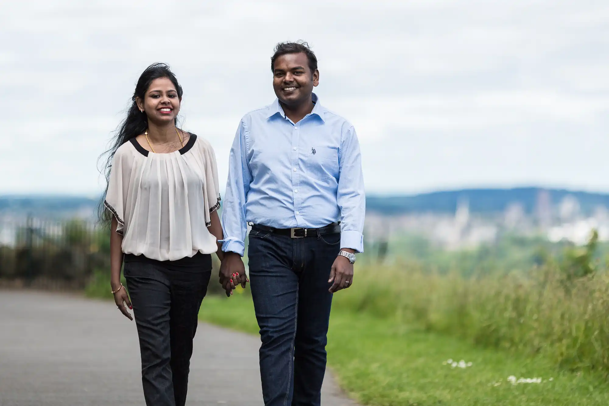 A man and a woman are walking outside on a pathway, holding hands and smiling. They are dressed in casual clothing, and there is greenery and a distant cityscape in the background.