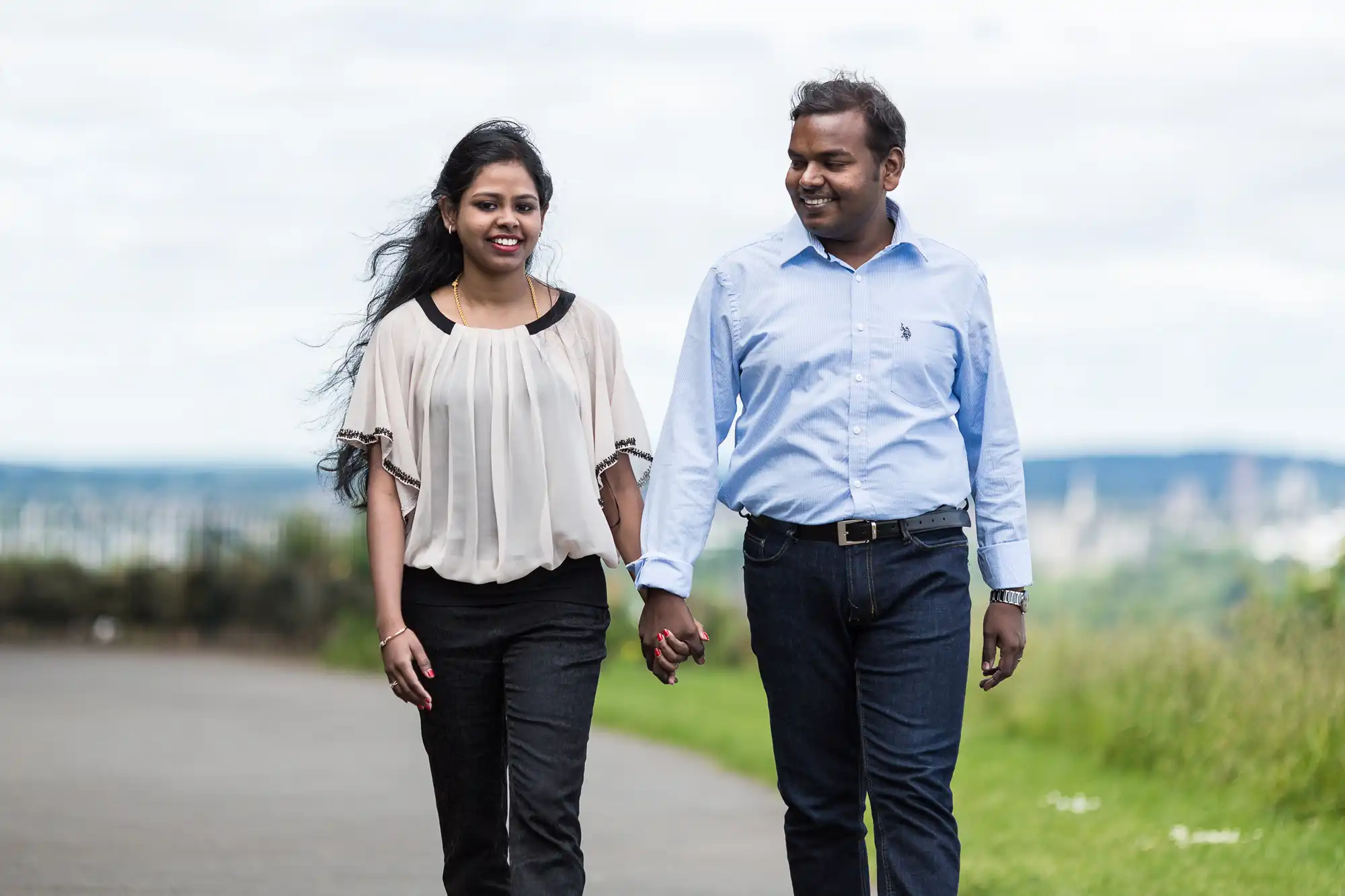 A couple walking outdoors on a paved path, holding hands. The woman has long hair and is wearing a light blouse, and the man is wearing a blue button-up shirt and dark pants.