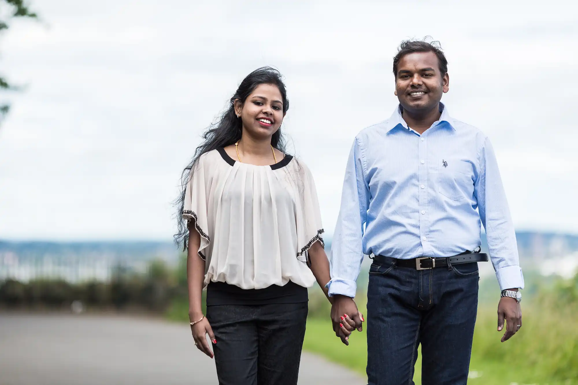 A couple, both casually dressed, walks hand in hand outdoors on a cloudy day. The woman wears a white blouse and black pants, while the man wears a light blue shirt and dark jeans.