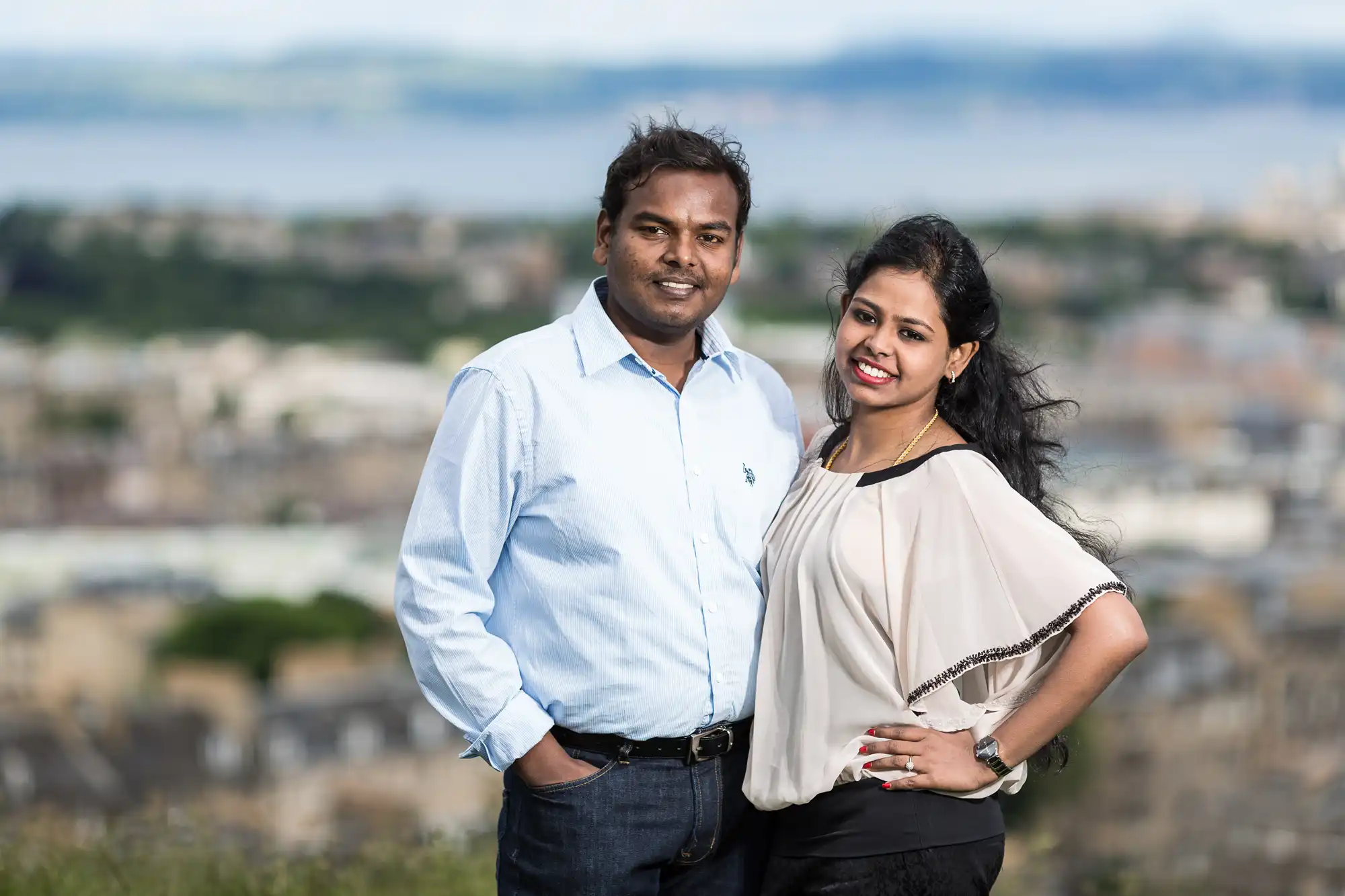 A man and woman stand side by side outdoors, smiling at the camera with a cityscape and distant water in the background.