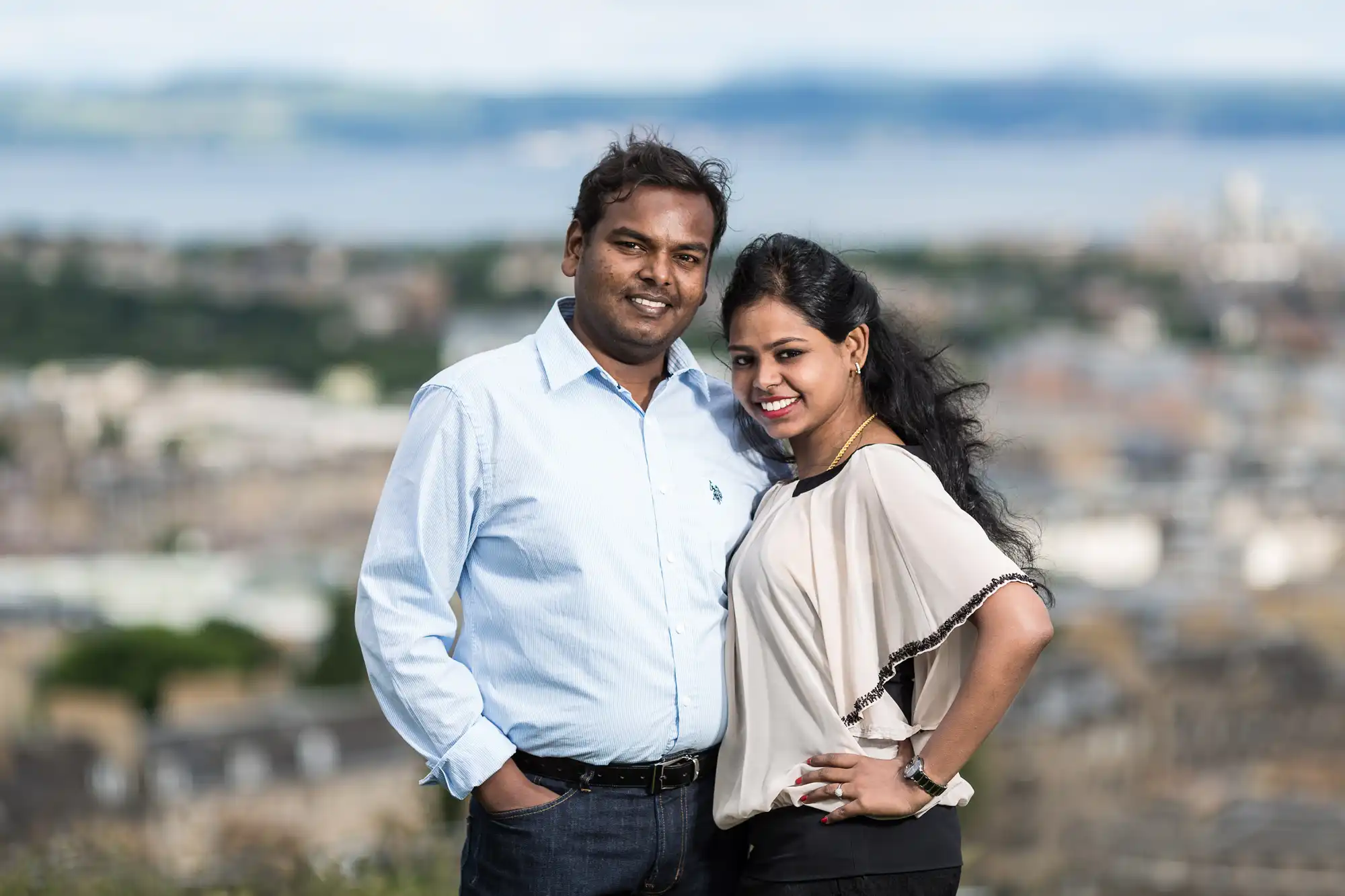 A man and woman stand close together, smiling at the camera with an expansive, blurred urban landscape in the background. Both are casually dressed and appear happy.