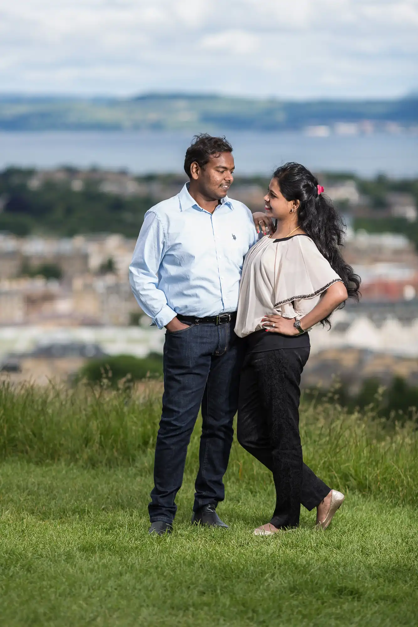 A man and a woman standing on a grassy area with a scenic landscape and body of water in the background, smiling at each other.