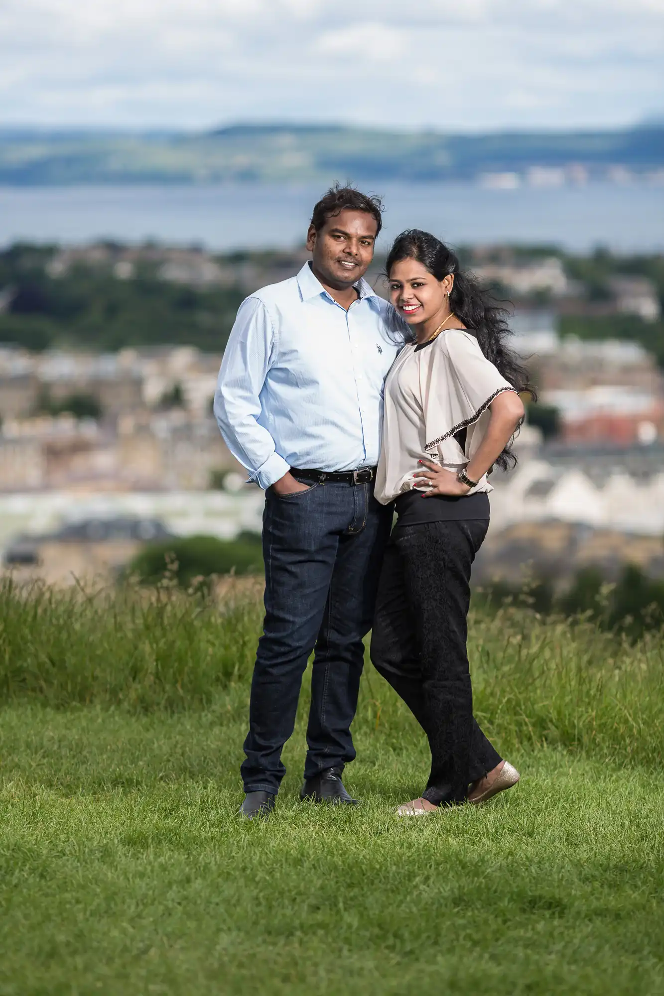 A man and woman stand together on a grassy area with a scenic view of a town and water in the background.