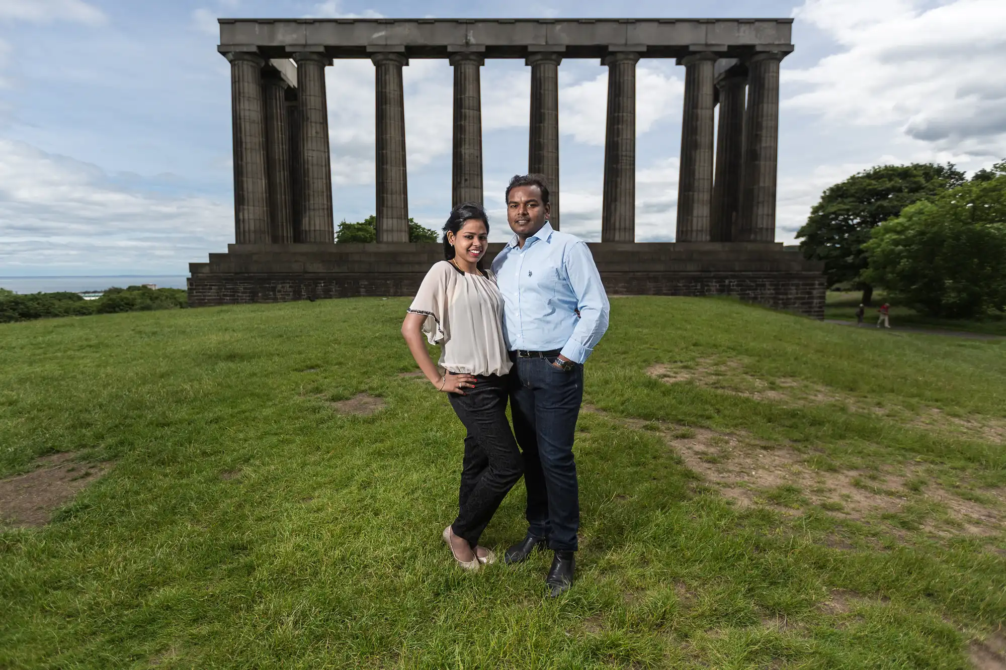 A man and woman stand on a grassy area in front of a structure with tall columns on a cloudy day.