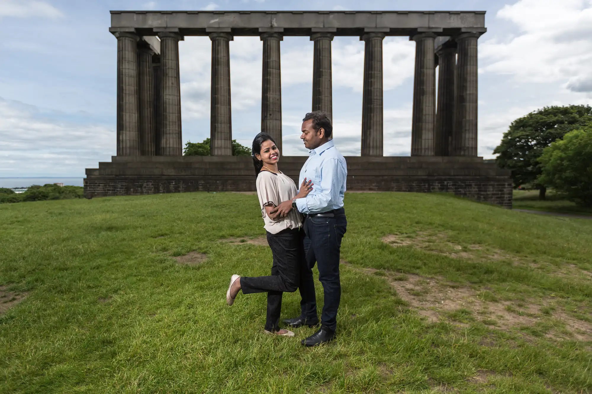 A couple stands closely together on a grassy area in front of a large, columned stone structure. The woman lifts one leg while smiling, and the man looks at her, holding her waist.