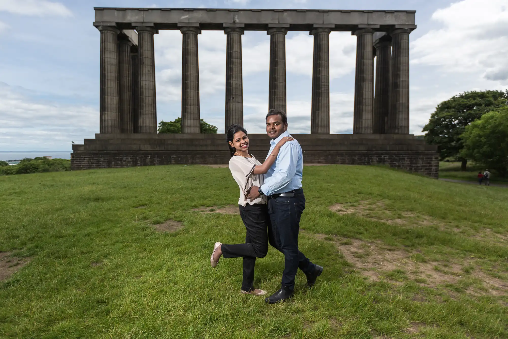 A couple embraces and smiles in front of a large stone structure with tall columns on a grassy area under a partly cloudy sky.