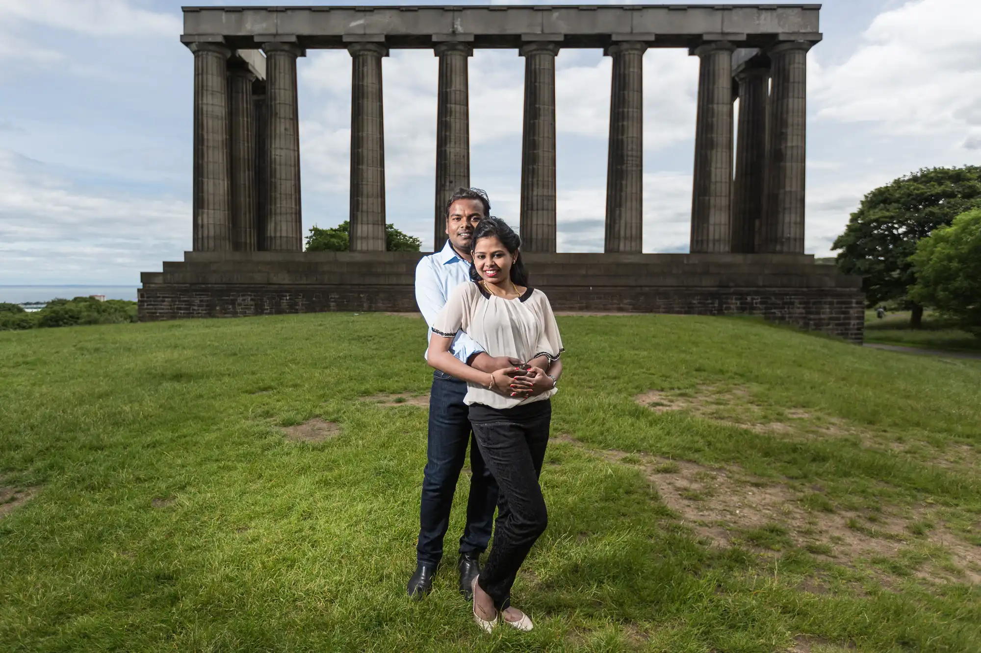 A couple stands closely together on a grassy area in front of a large neoclassical structure with columns on a partly cloudy day.