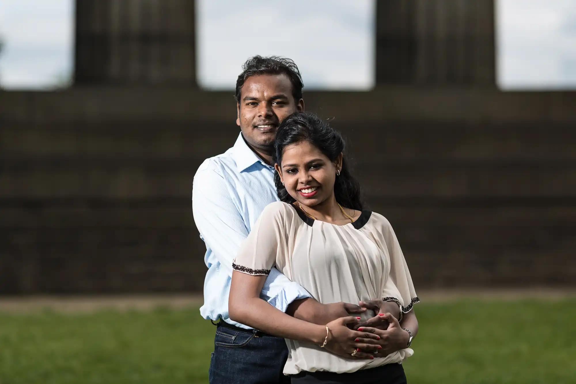 A couple stands outdoors in front of a stone structure, smiling and embracing. The man wears a light blue shirt, and the woman wears a white blouse.