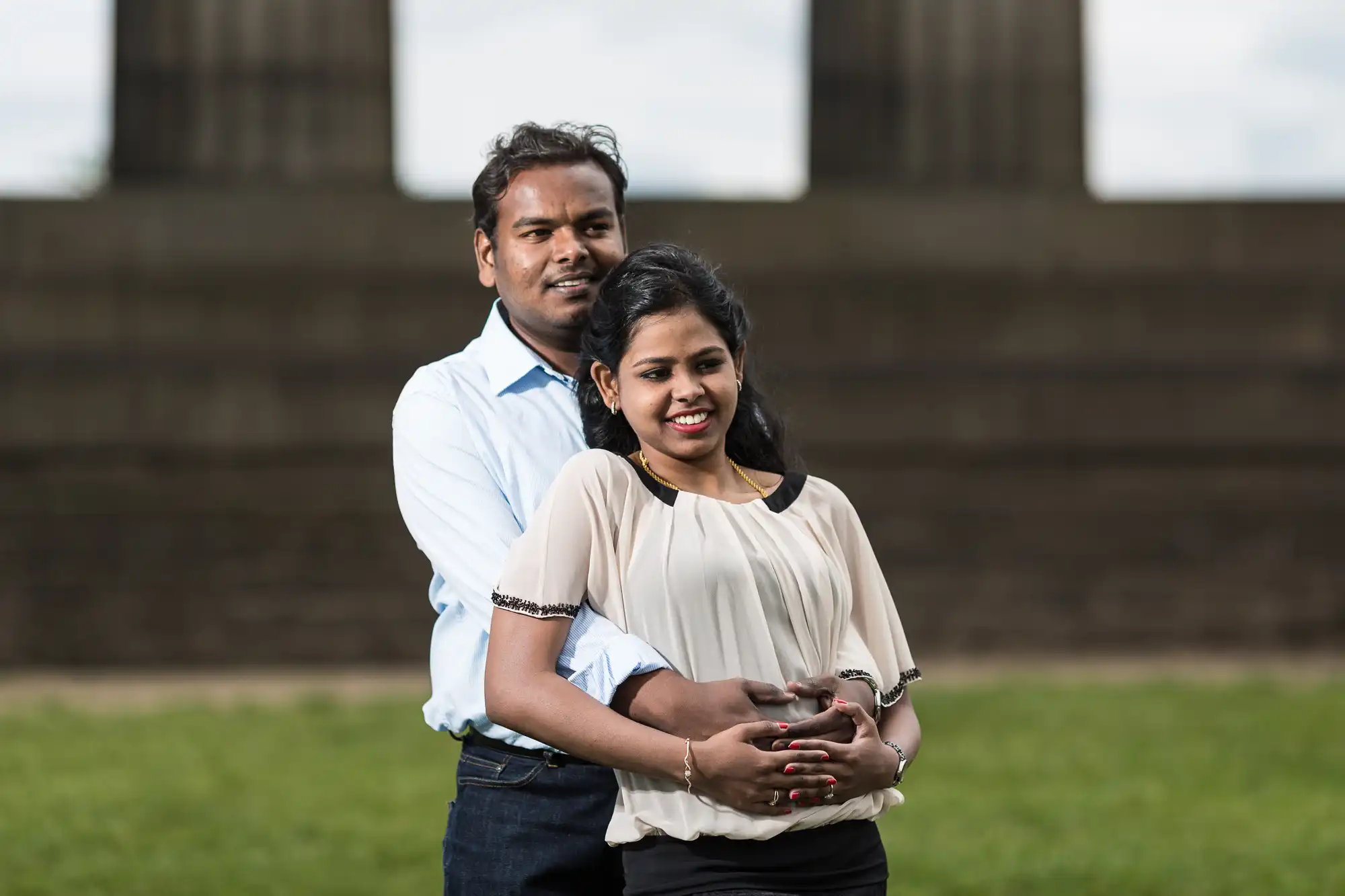 A man stands behind a woman, hugging her, as they both smile outdoors with a stone structure and green grass in the background.