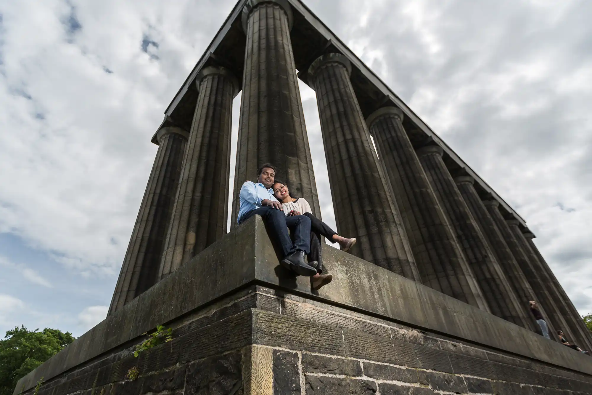 Two people sit on the stone base of a neoclassical monument with tall columns, beneath a partly cloudy sky.