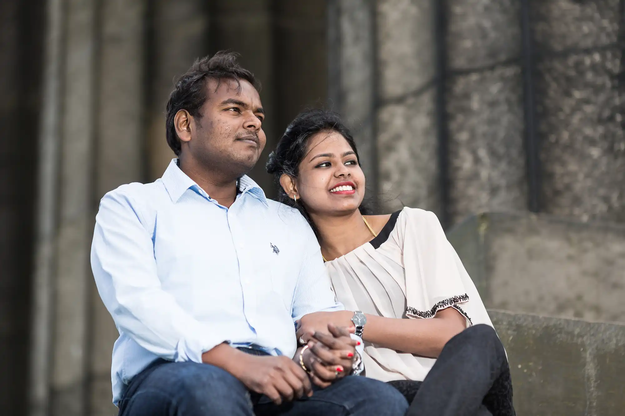 A man and a woman sit together on stone steps. The woman is resting her head on the man's shoulder and smiling. They look relaxed and content.