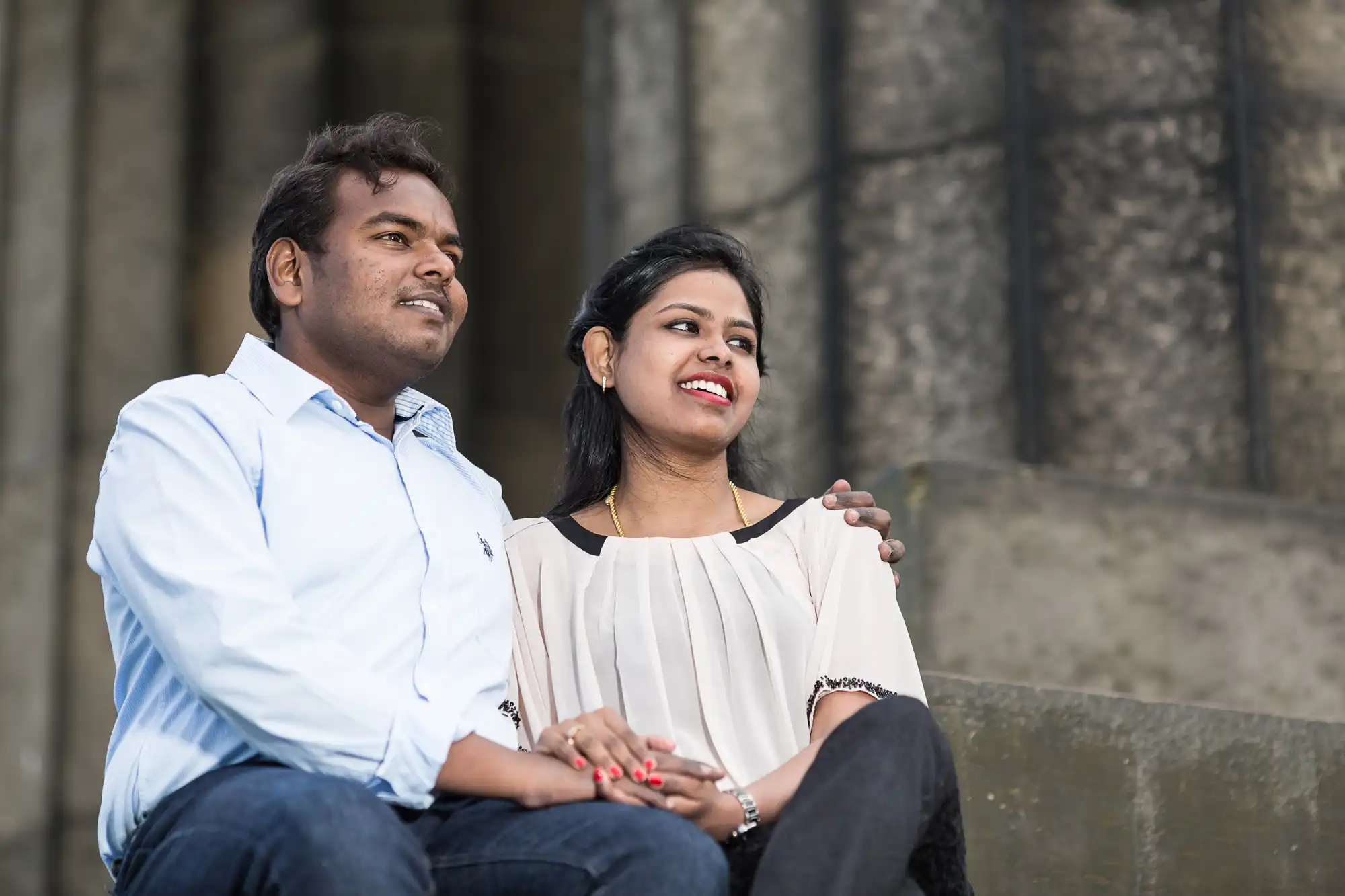 A man and a woman sit closely together on concrete stairs with the man's arm around the woman's shoulder. Both are smiling and looking at something off-camera.