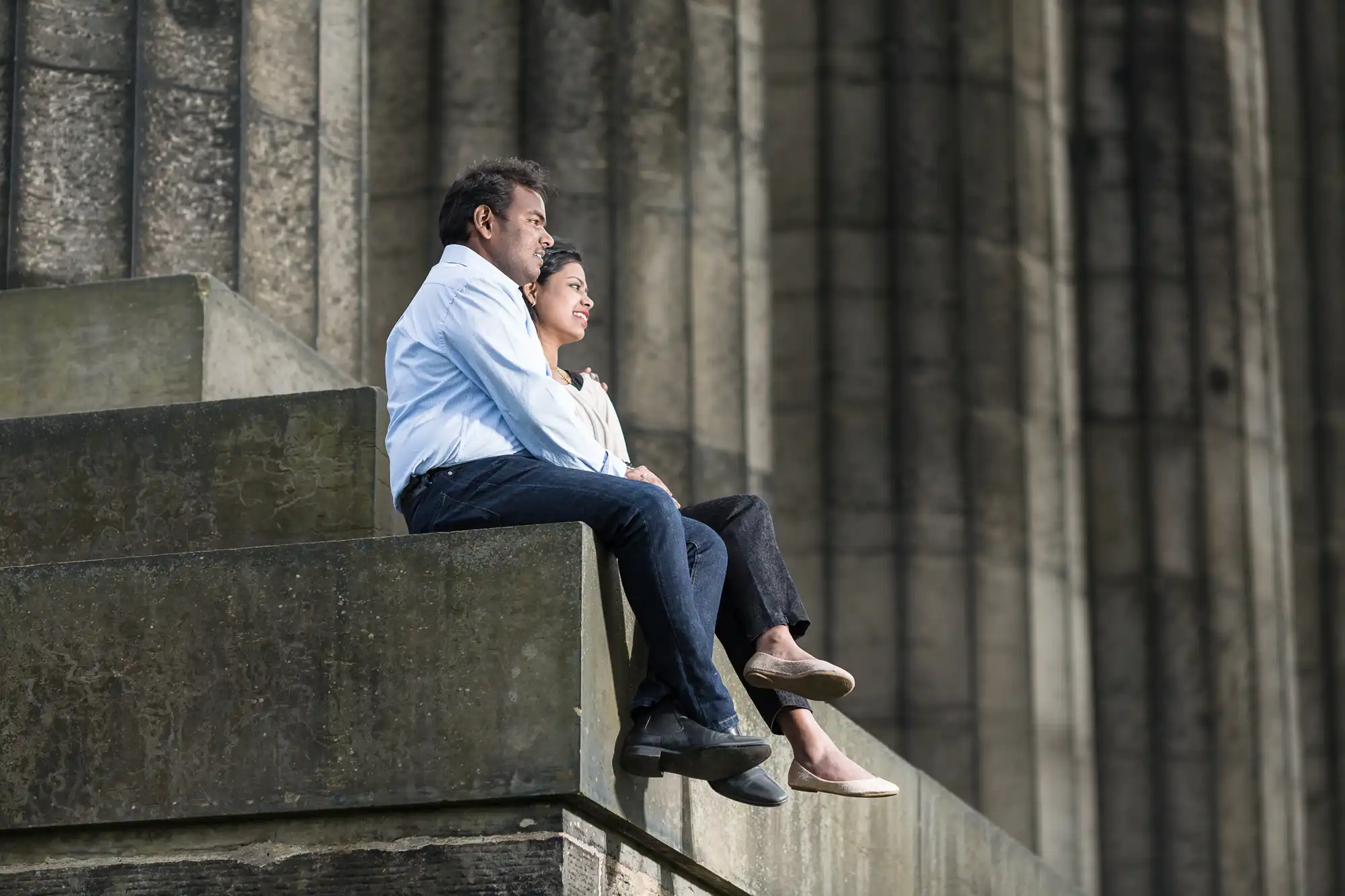 A man and a woman sit on a concrete structure, facing forward. The background consists of large, vertical stone pillars. Both are casually dressed and appear to be in a relaxed state.