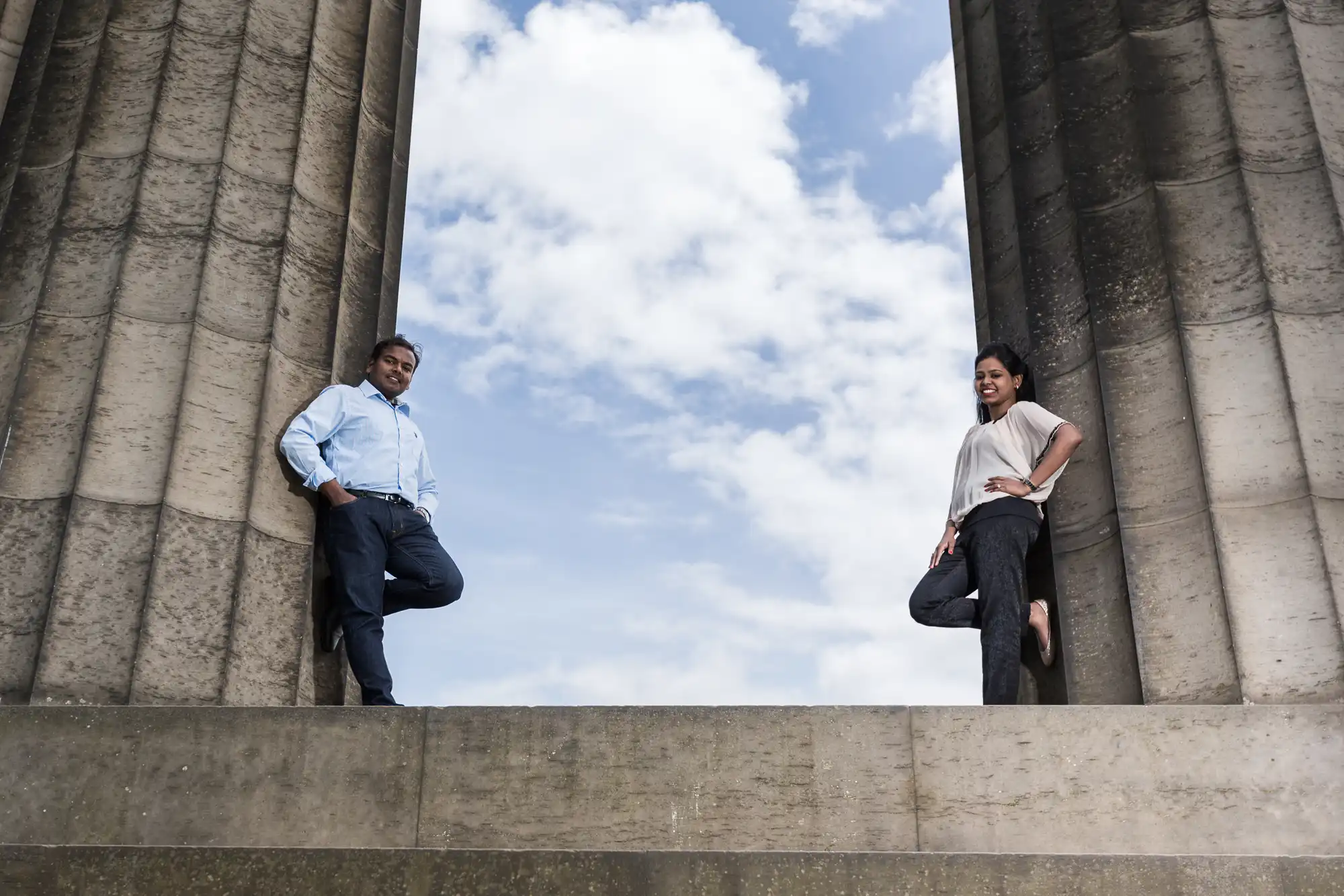 Two people stand on a ledge between two large stone columns, posing against a backdrop of a partly cloudy sky.