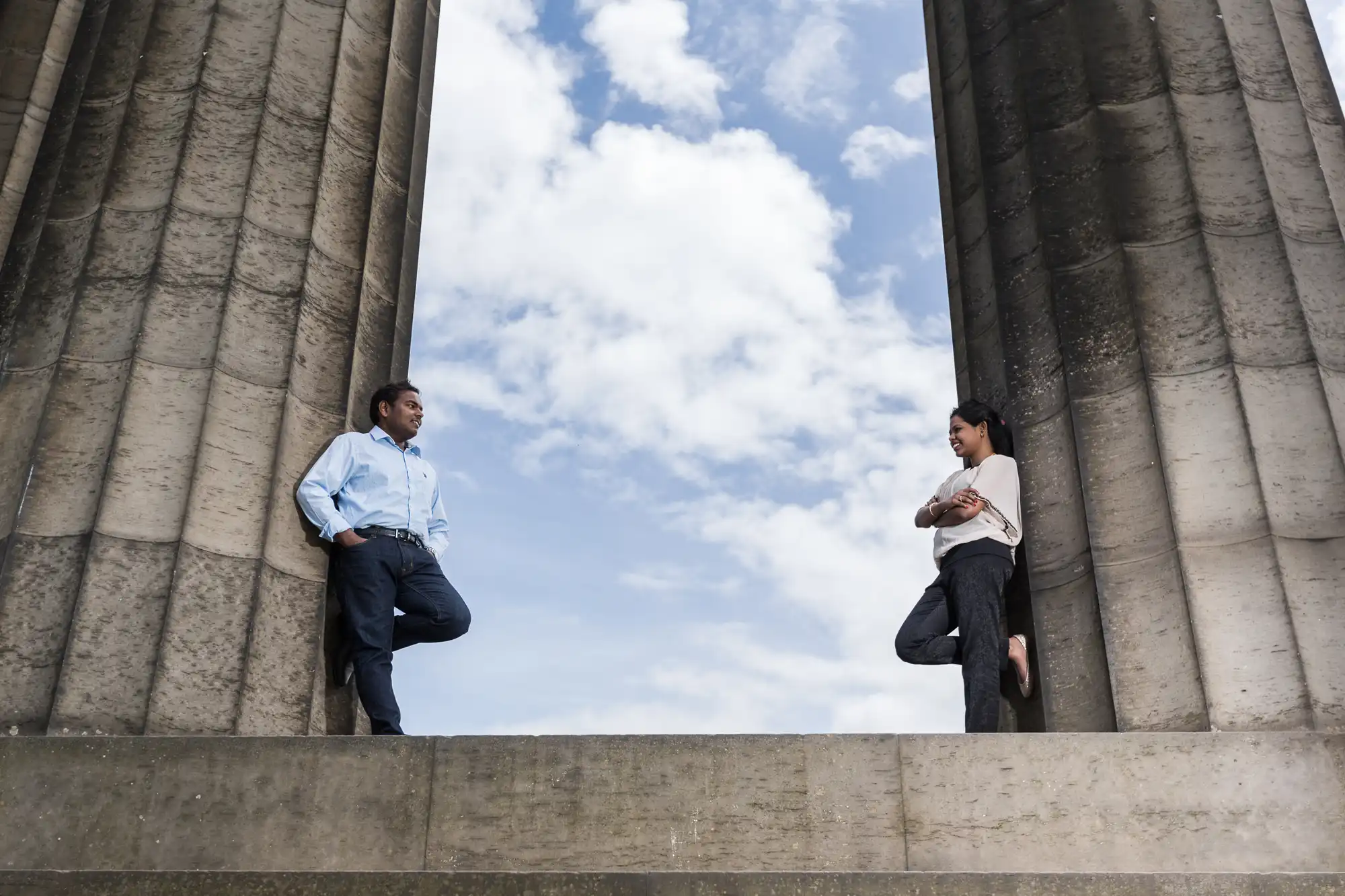 Two people stand facing each other while leaning against tall stone pillars with a cloudy sky in the background.