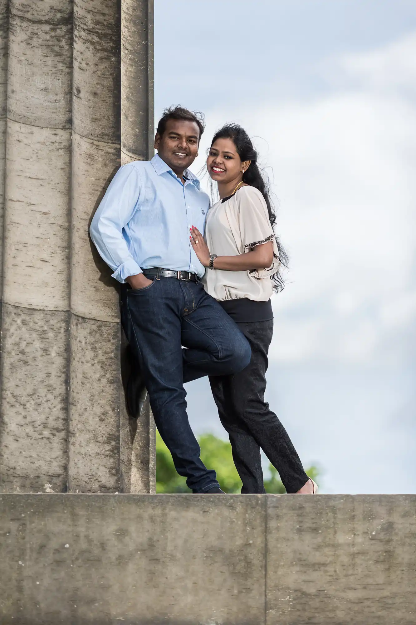 A man and a woman smile while posing leaning against a stone column. The man is wearing a blue shirt and jeans, and the woman is wearing a light blouse and dark pants.