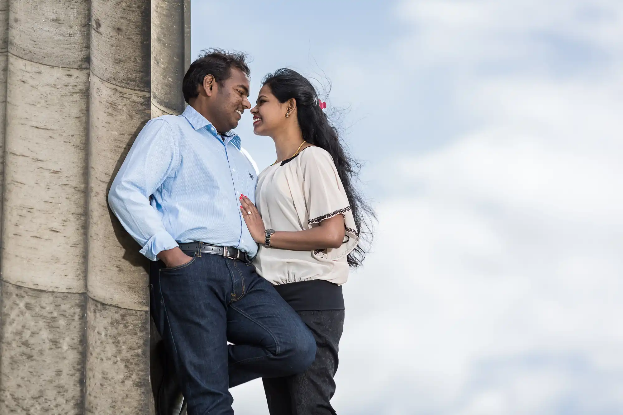 A man and a woman stand close to each other outdoors, leaning against a stone structure. They are smiling and looking at each other affectionately. The sky is cloudy in the background.