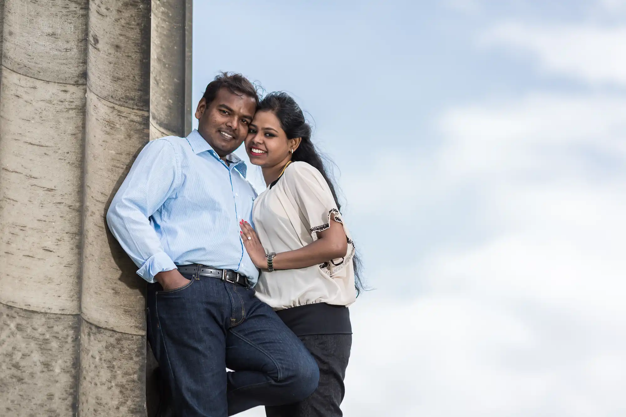 A man and woman lean against a stone structure, smiling at the camera on a partly cloudy day.