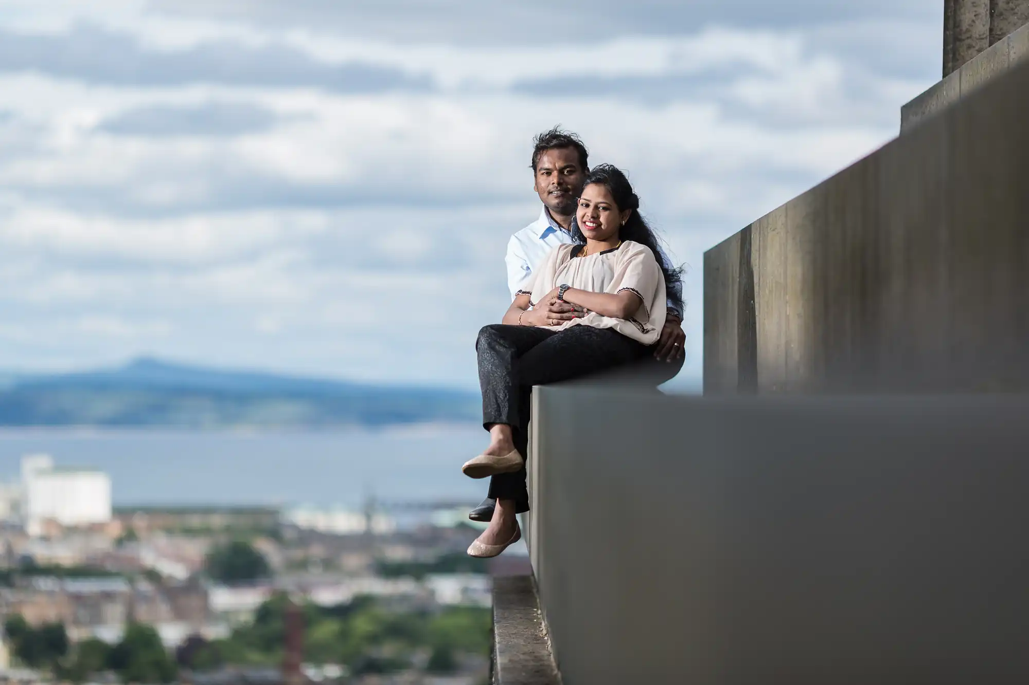 A couple sits together on a ledge with a cityscape and body of water in the background under a cloudy sky.