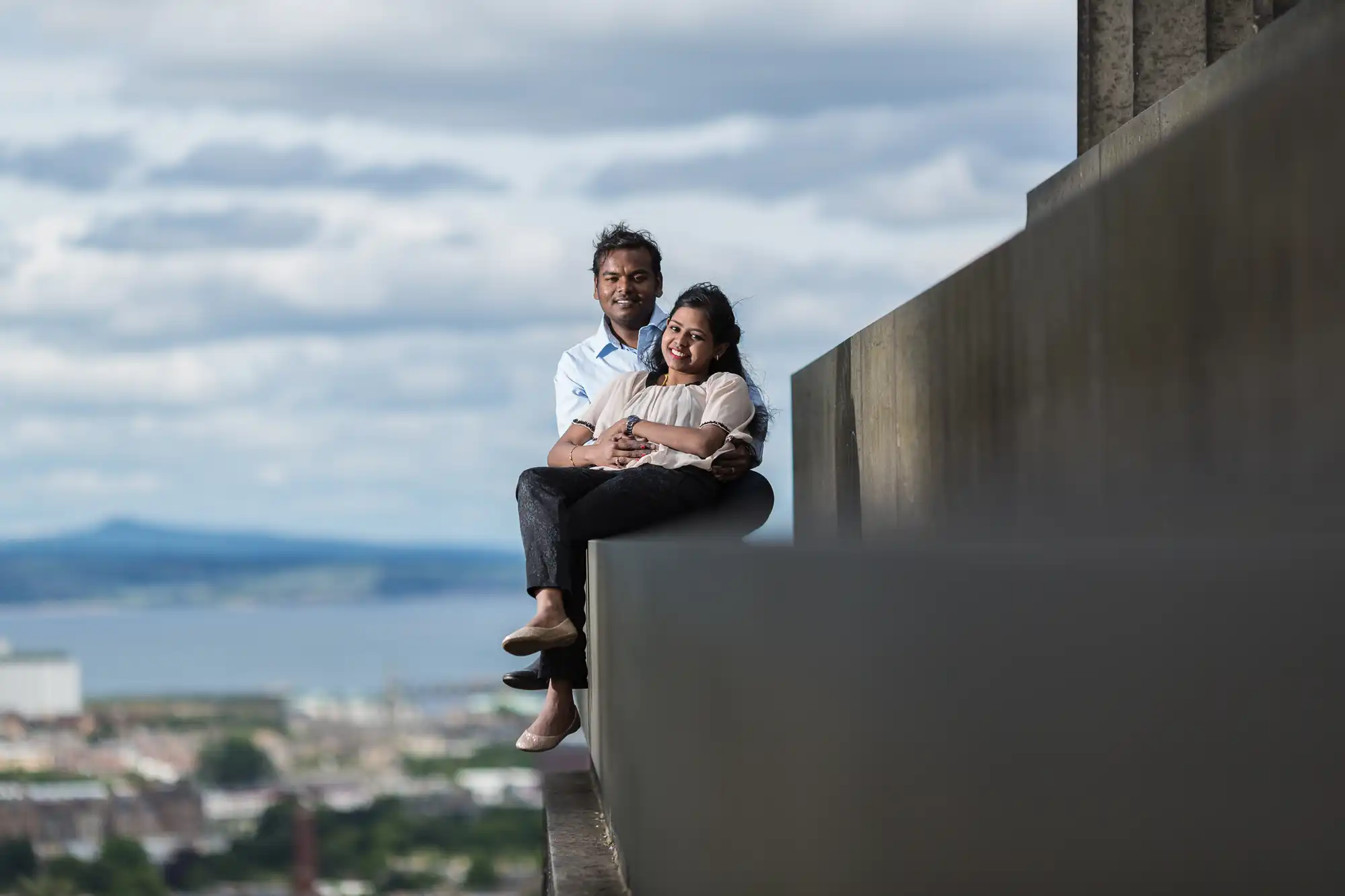 A man and a woman sit closely, smiling, on a ledge overlooking an expansive view of a city and landscape under a cloudy sky.