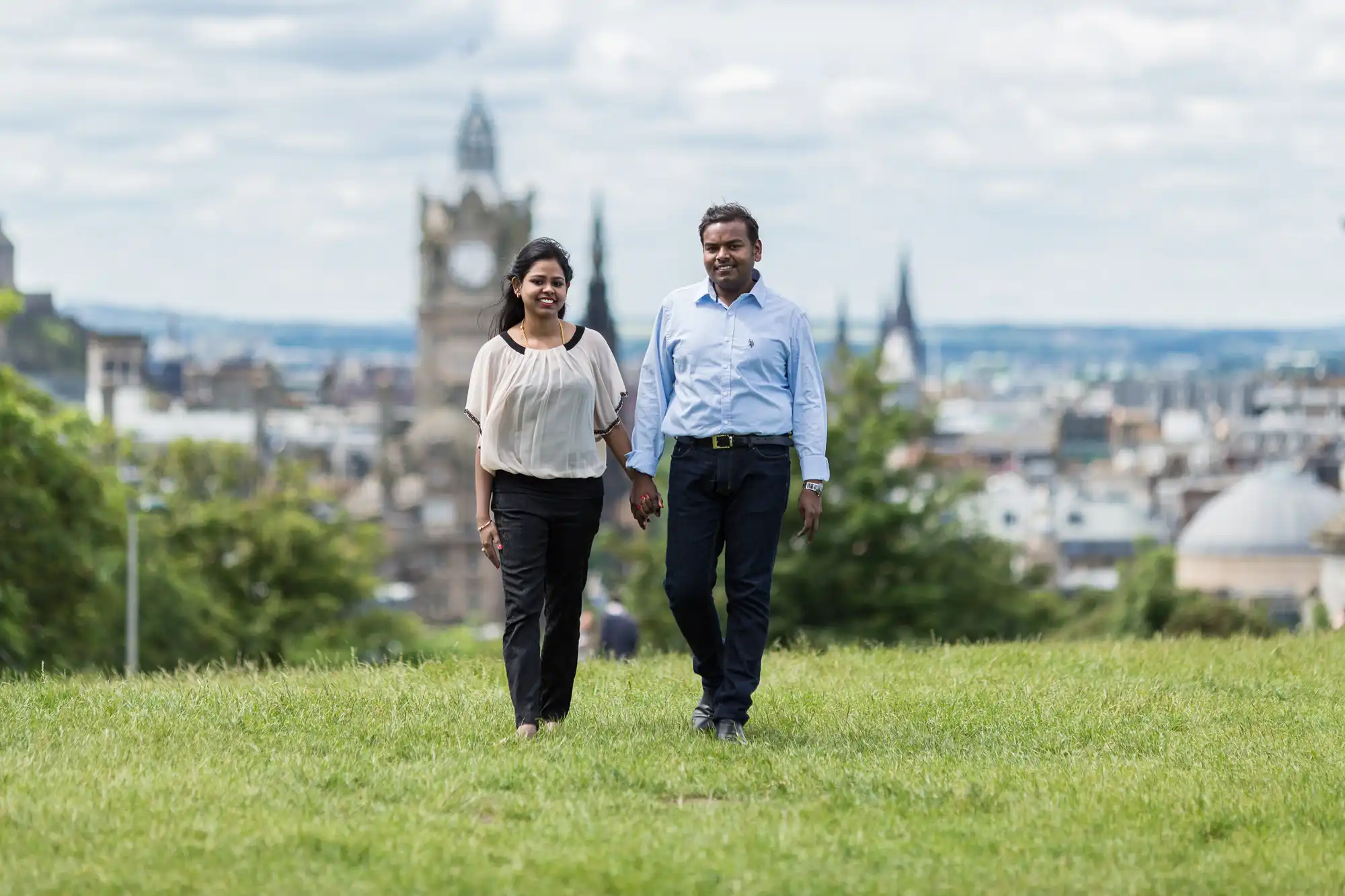 A man and a woman holding hands, walking on a grassy area with a cityscape and clock tower in the background.