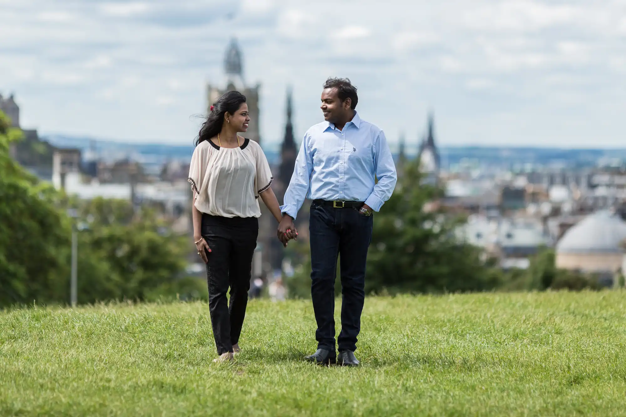 A couple holding hands while walking on a grassy area with a cityscape and cloudy sky in the background.