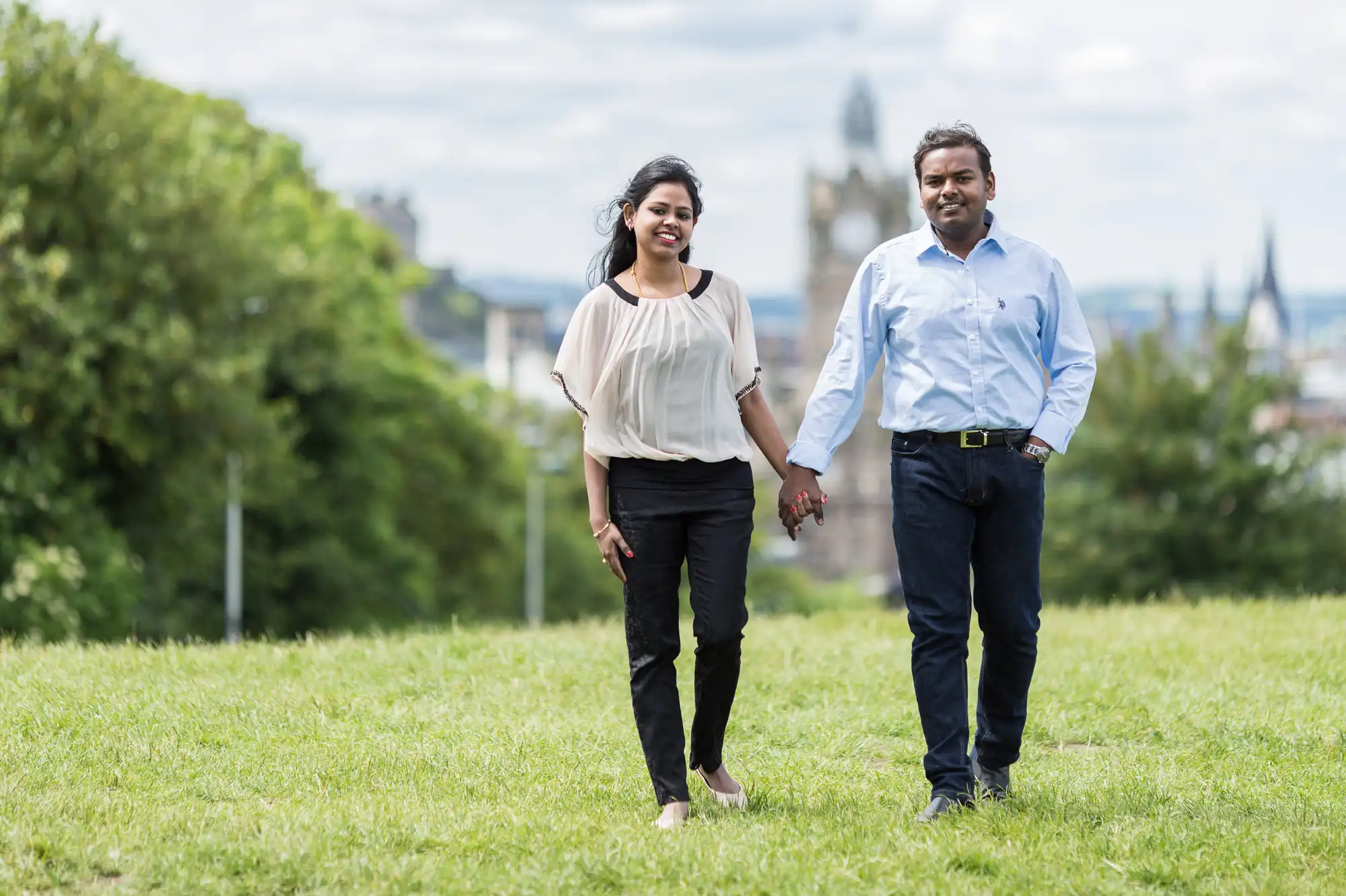 A man and a woman holding hands walk on a grassy field with trees and a cityscape in the background.