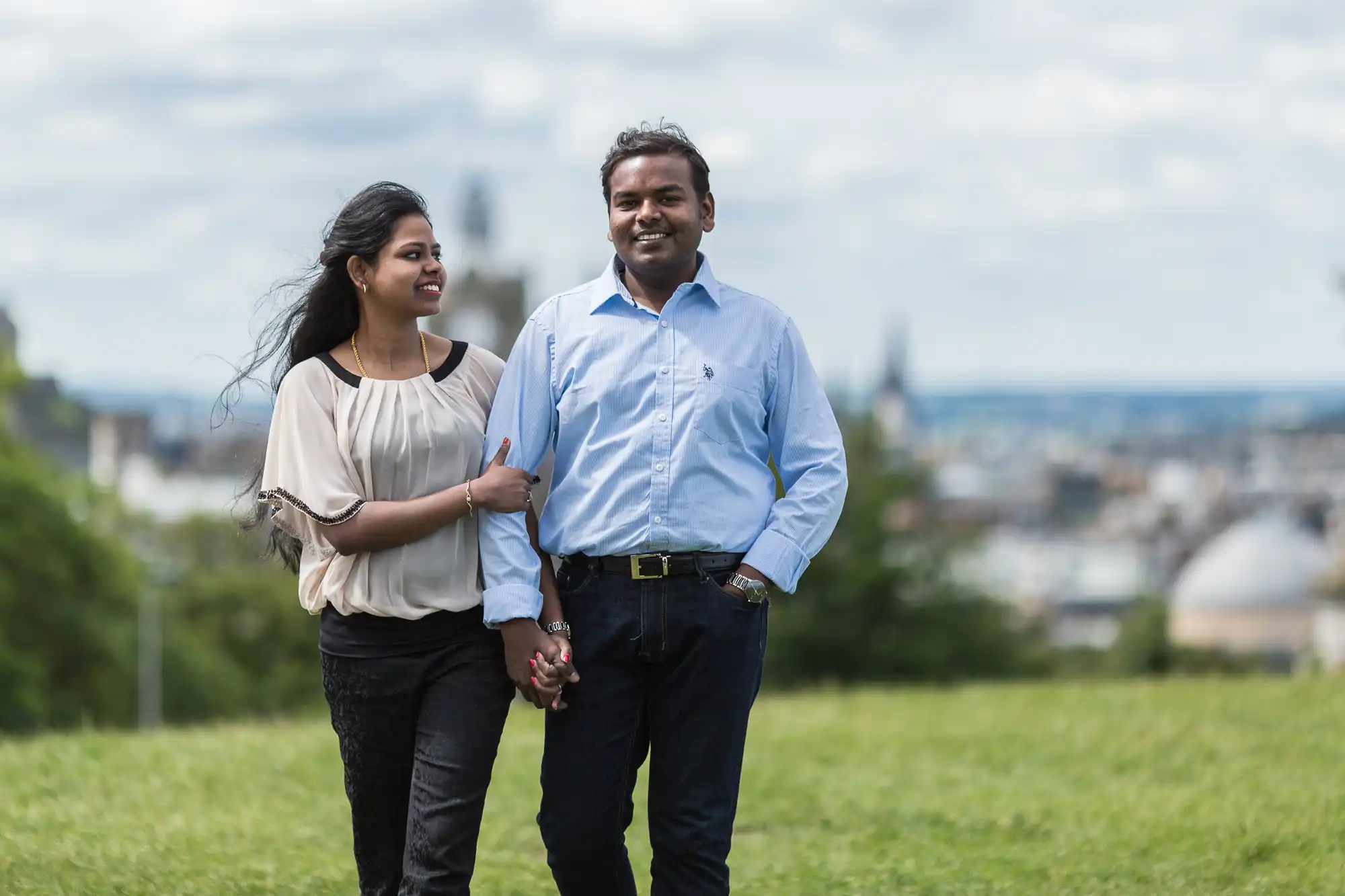 A couple is walking hand in hand on a grassy area, with a cityscape in the background. They both appear happy and relaxed.