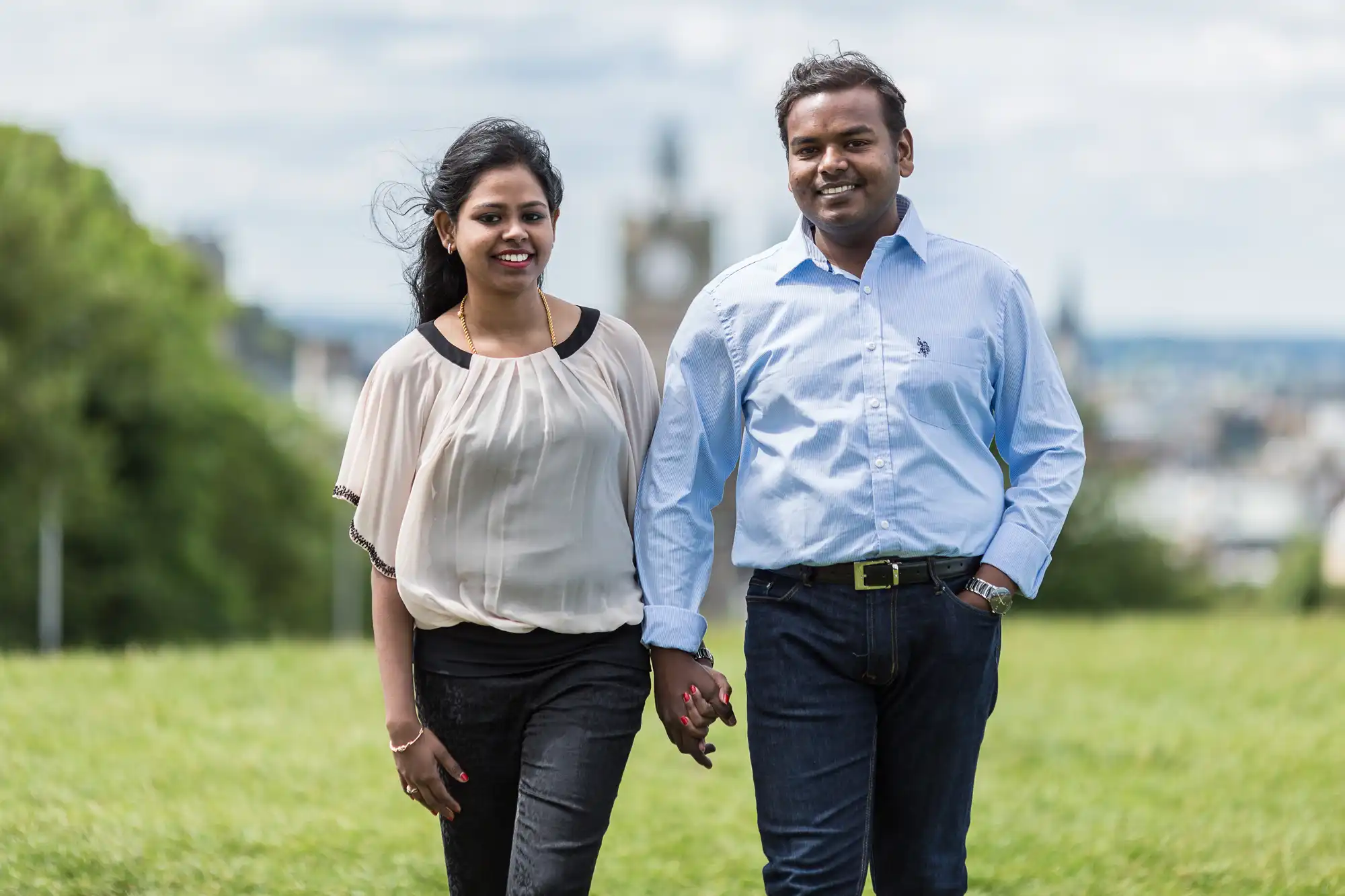 A couple holds hands while walking on a grassy field, both smiling. The man wears a blue shirt and black pants, and the woman wears a light blouse and black pants. Trees and a distant cityscape are in the background.