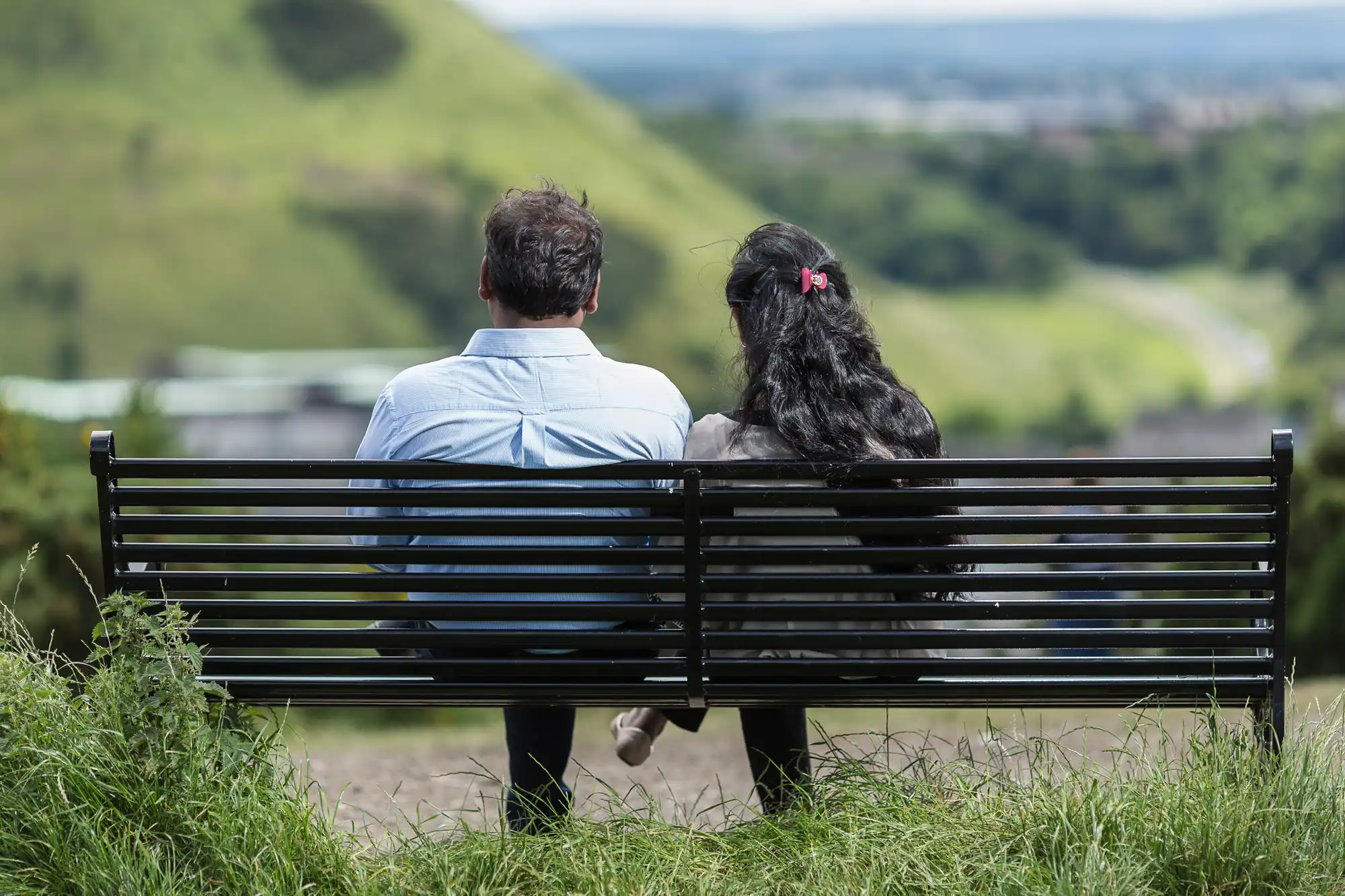 Two people sit on a black bench overlooking a scenic, green landscape.