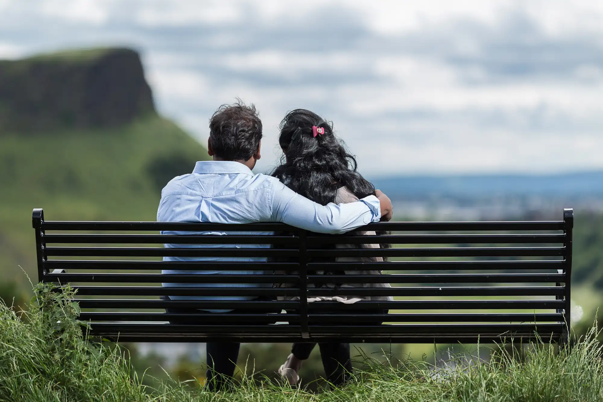 A man and a woman sit on a bench, with the man’s arm around the woman, facing a scenic landscape with hills and cloudy skies in the background.