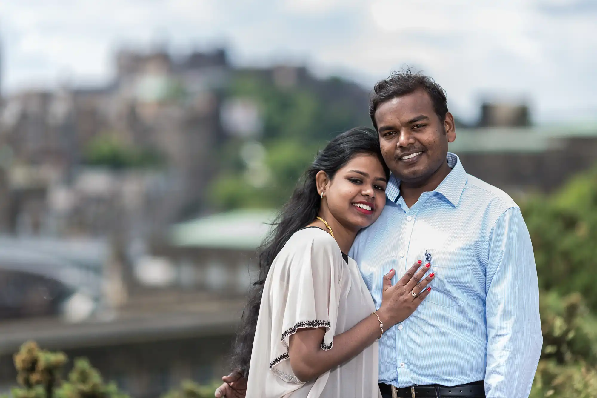 A couple stands closely together outdoors, both smiling at the camera. The background is blurred, displaying a natural setting with a structure in the distance.