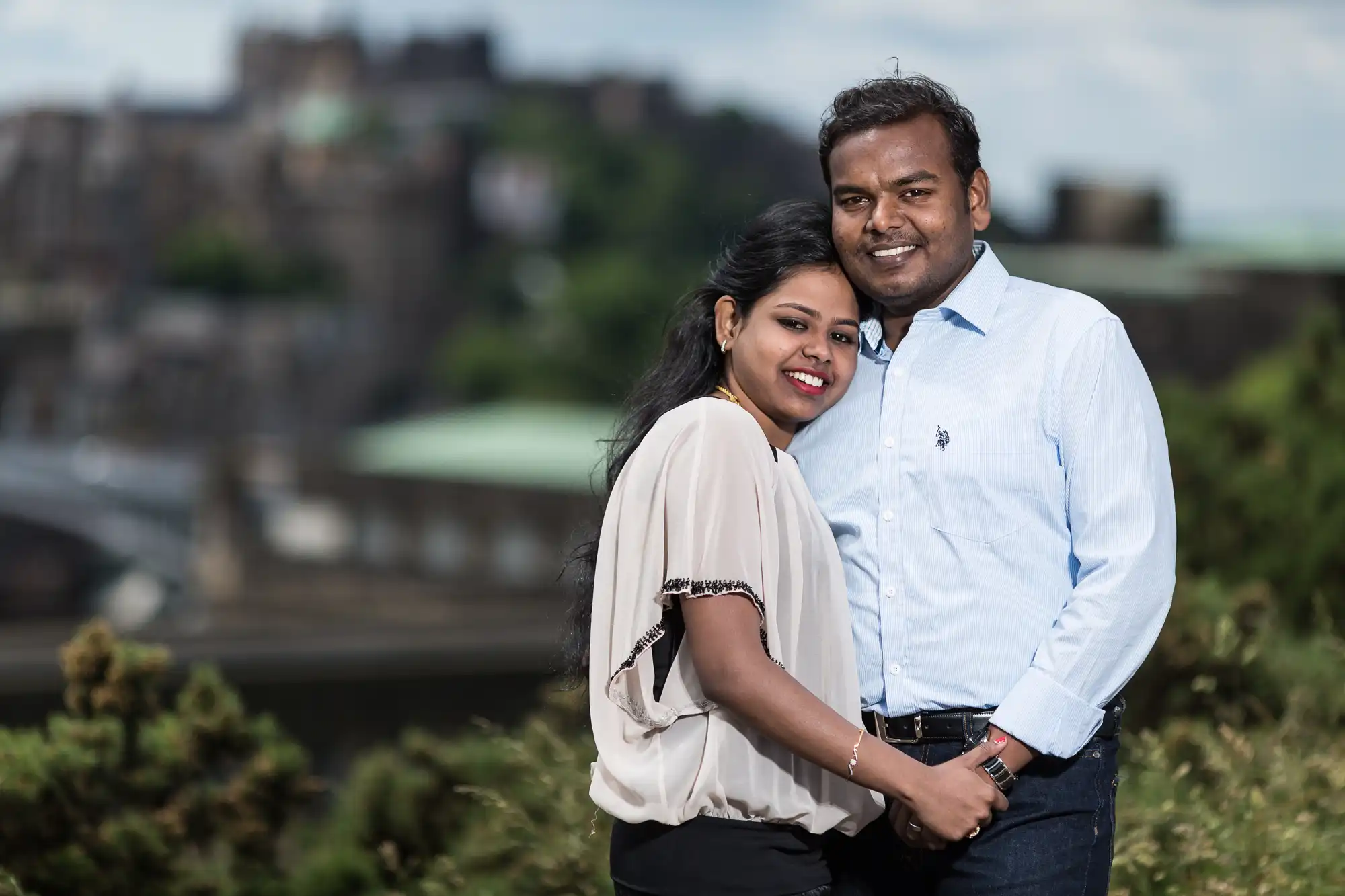 A couple stands closely together outdoors in front of a blurred scenic background. The woman wears a light blouse and black pants, while the man wears a light blue shirt and dark pants. They are both smiling.