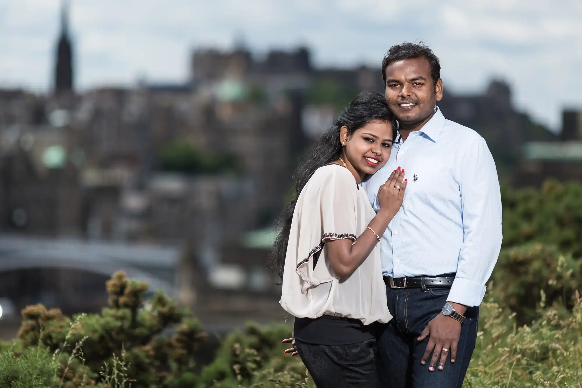 A couple stands together outdoors, smiling, with a blurred cityscape and greenery in the background.