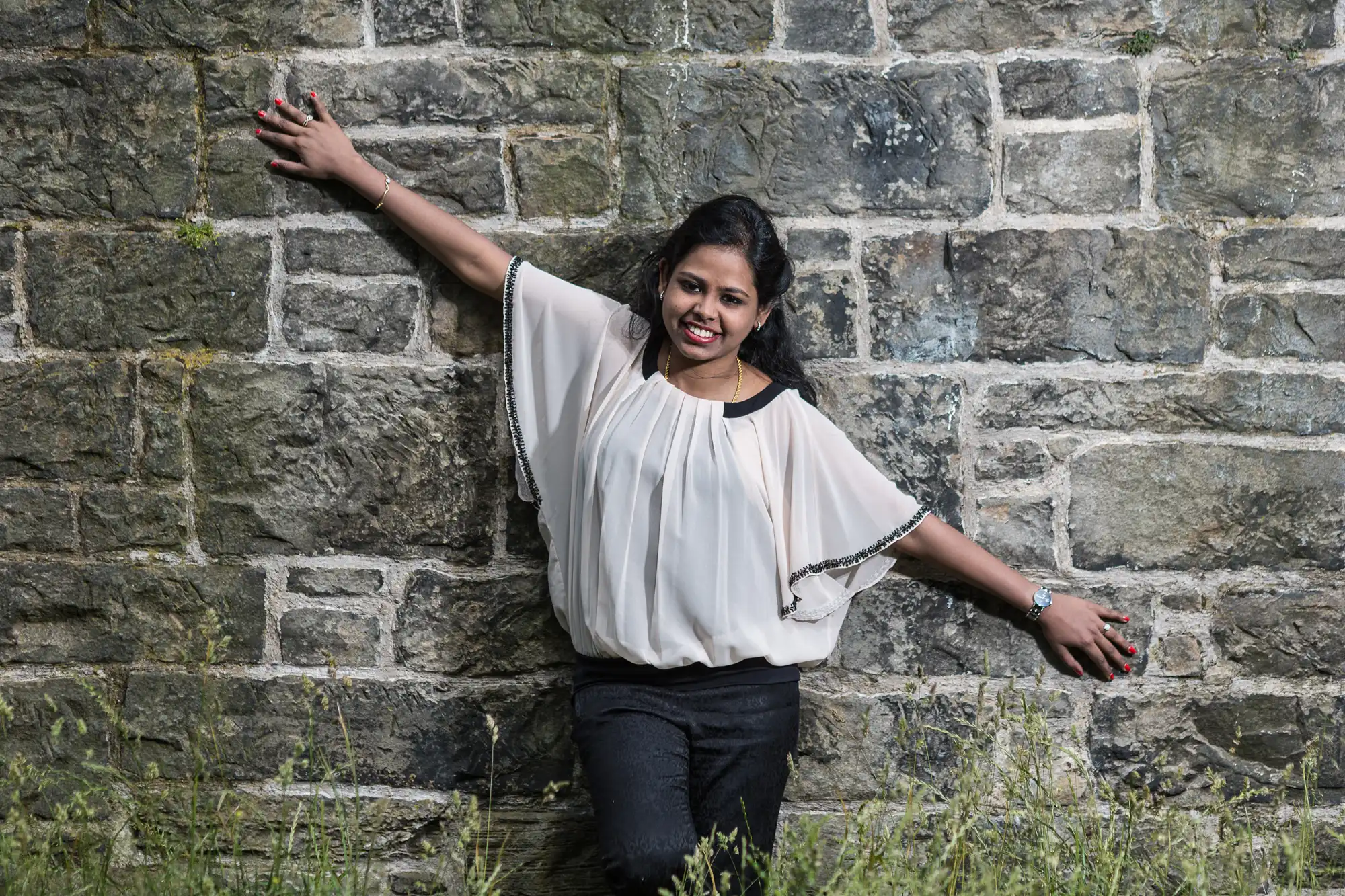 A woman smiles while leaning against a stone wall with her arms outstretched. She is wearing a white blouse and black pants.