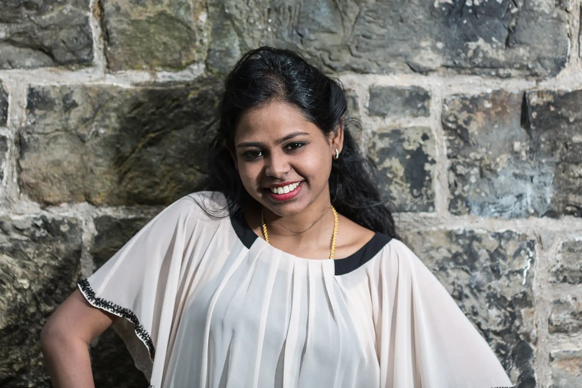 A woman with long black hair and a white blouse poses in front of a stone wall, smiling at the camera.