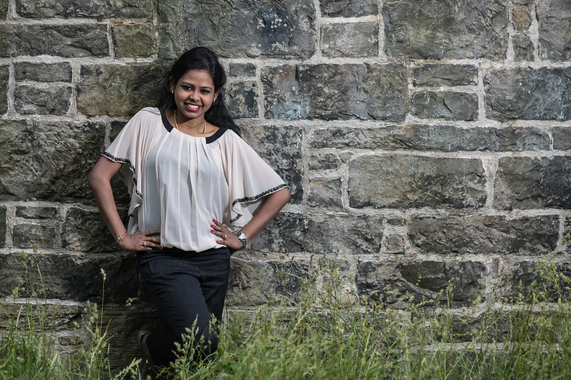 A woman stands smiling with hands on hips, leaning against a weathered stone wall. She is wearing a light-colored top and dark pants. Grass grows at the base of the wall.