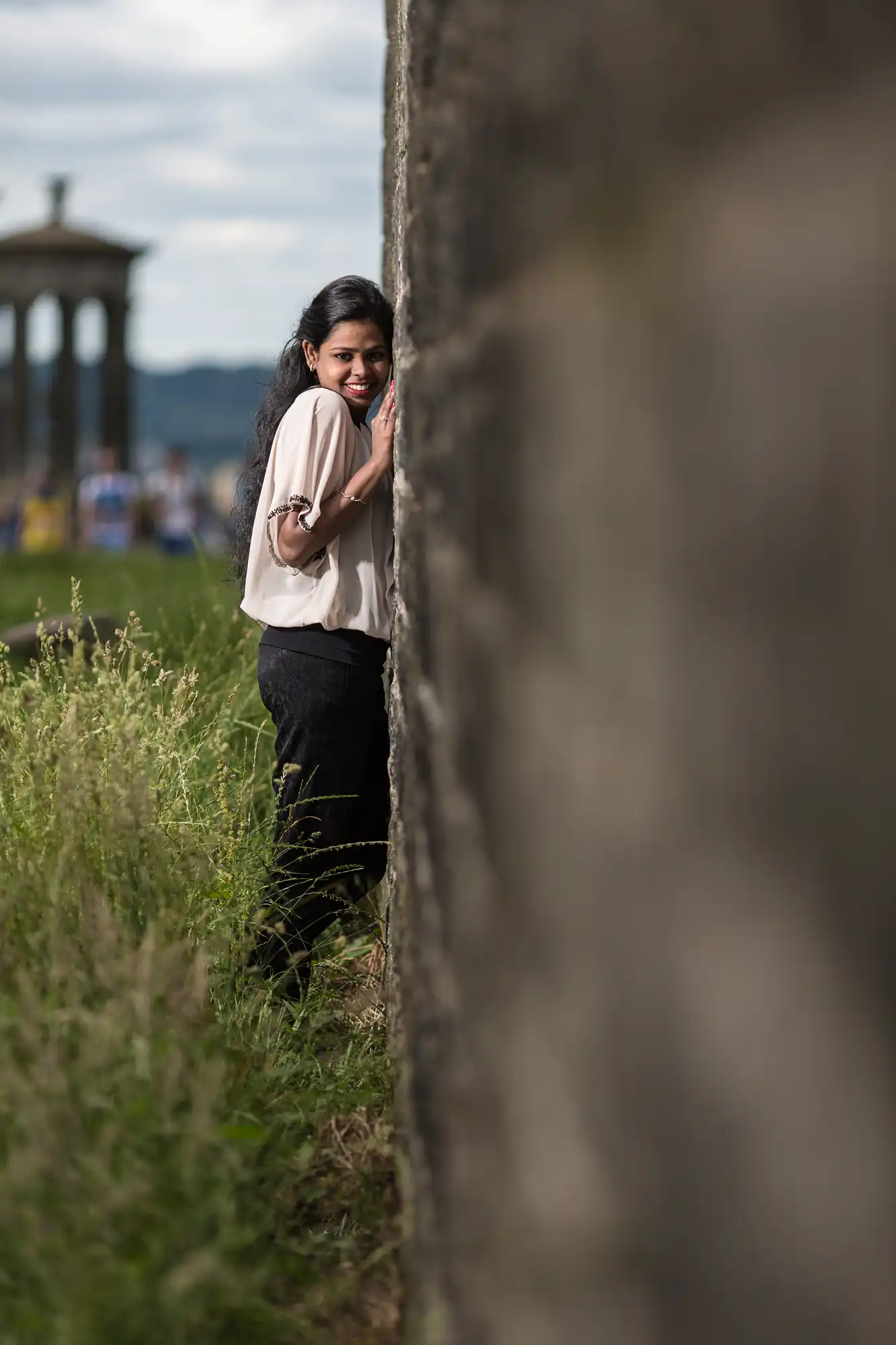 A woman with long dark hair leans against a stone wall outdoors, partially hidden by the wall. She is wearing a light blouse and dark pants, with a structure visible in the background.