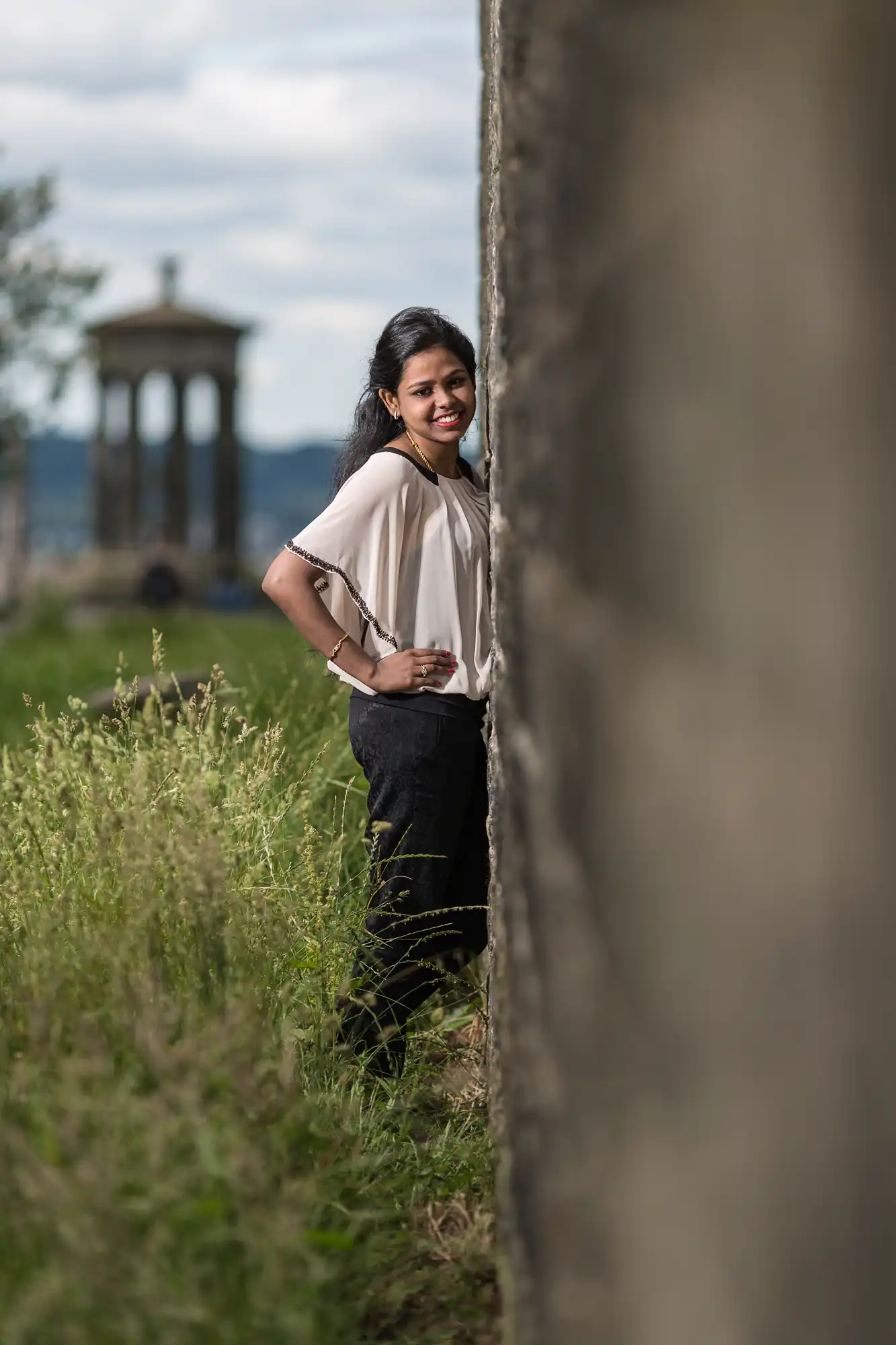 A woman stands and smiles next to a stone wall with a historical monument blurred in the background.