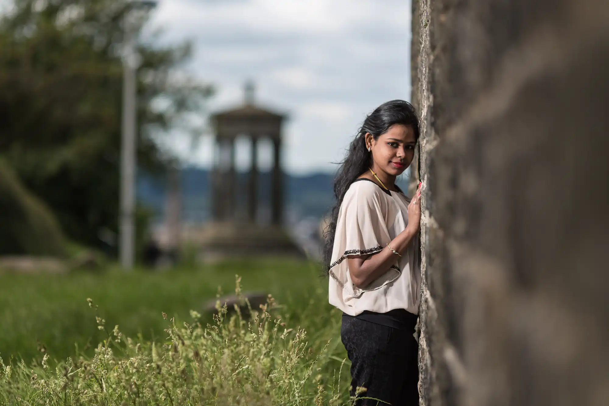 A woman stands outdoors, leaning against a stone wall, with a grassy area and a structure in the background.