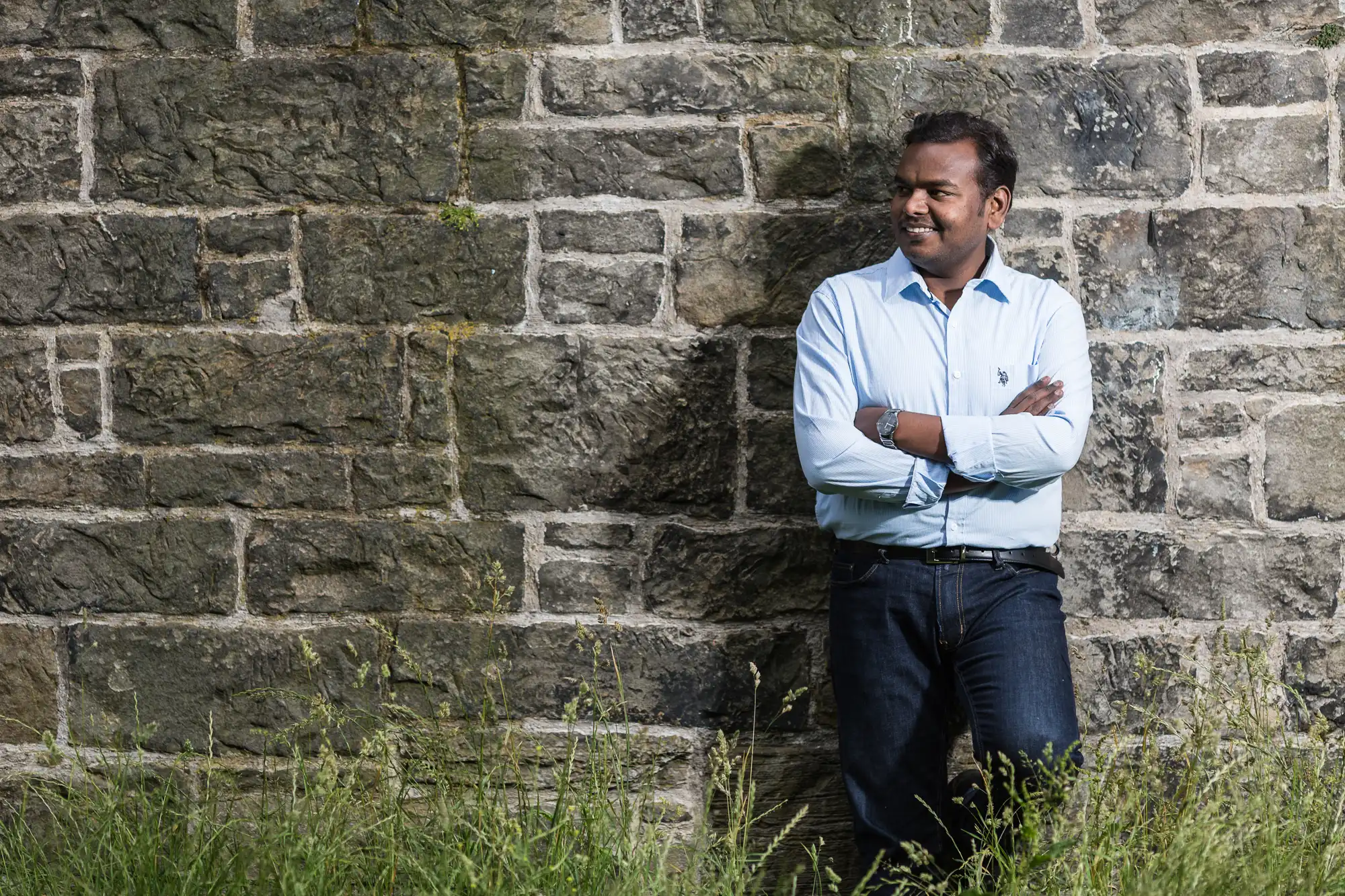 Man in a blue shirt stands with arms crossed, leaning against a stone wall with grass in the foreground.