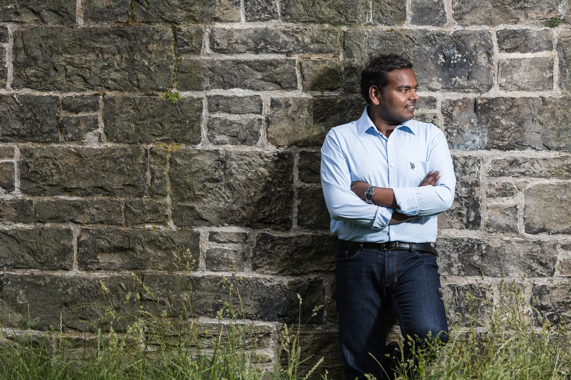 A man in a light blue shirt and dark jeans stands with arms crossed leaning against a stone wall, with grass in the foreground.