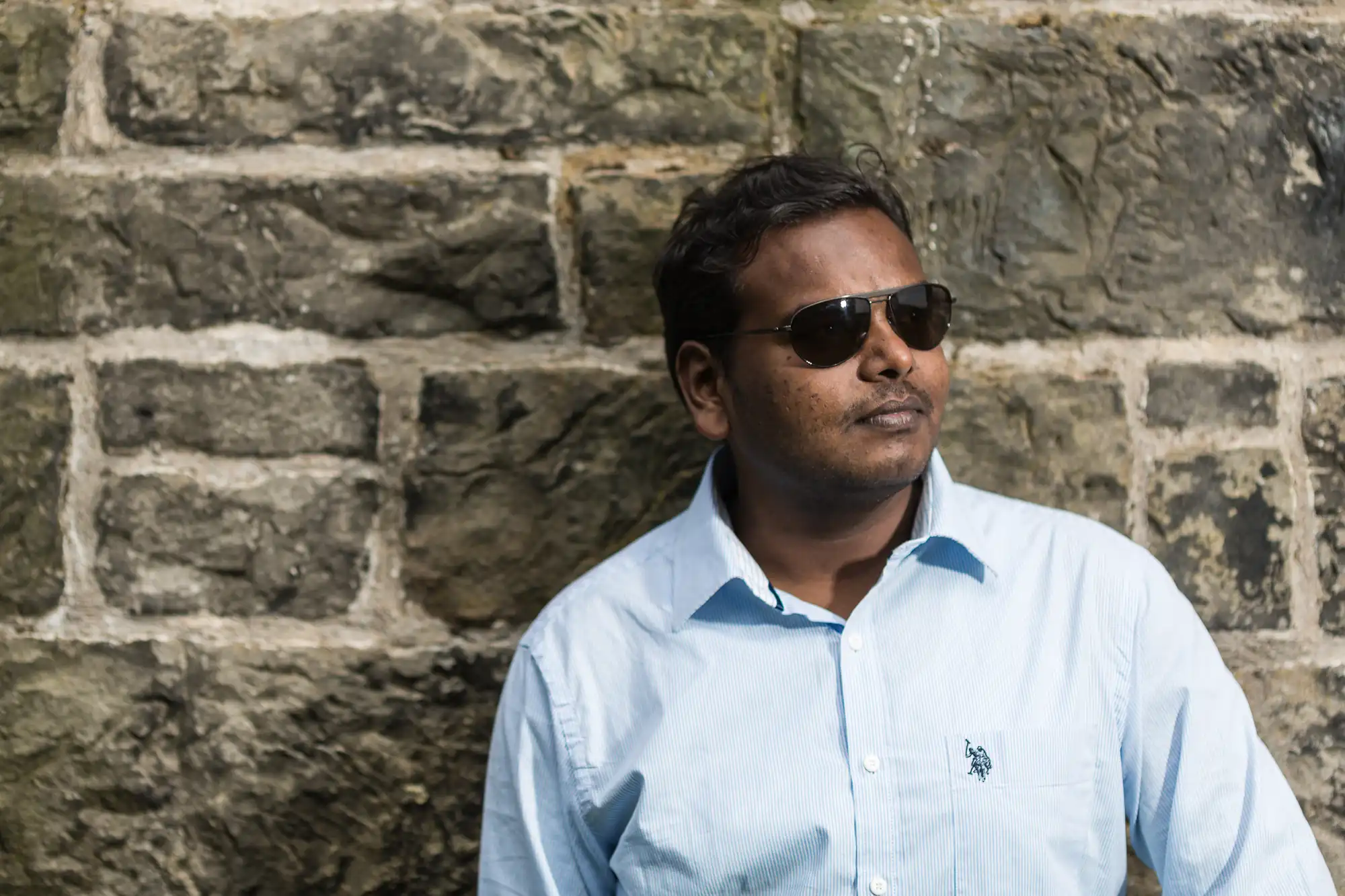 A person wearing sunglasses and a light blue shirt stands in front of a stone wall, looking off into the distance.