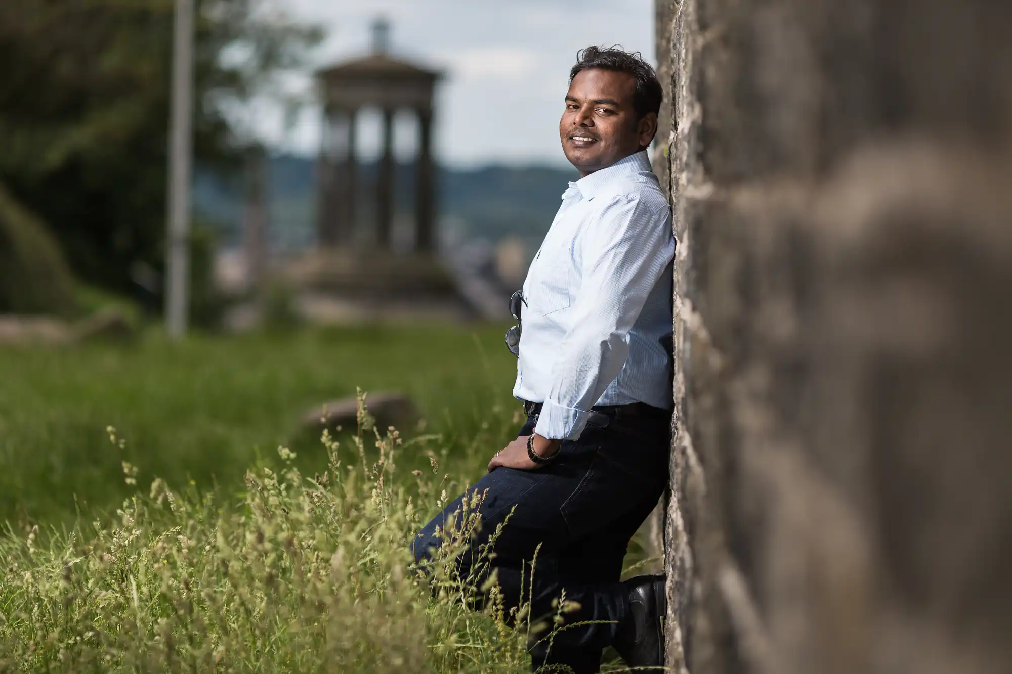 Man in a white shirt and dark pants leans against a stone wall outdoors, with a blurred ancient structure in the background and tall grass surrounding him.