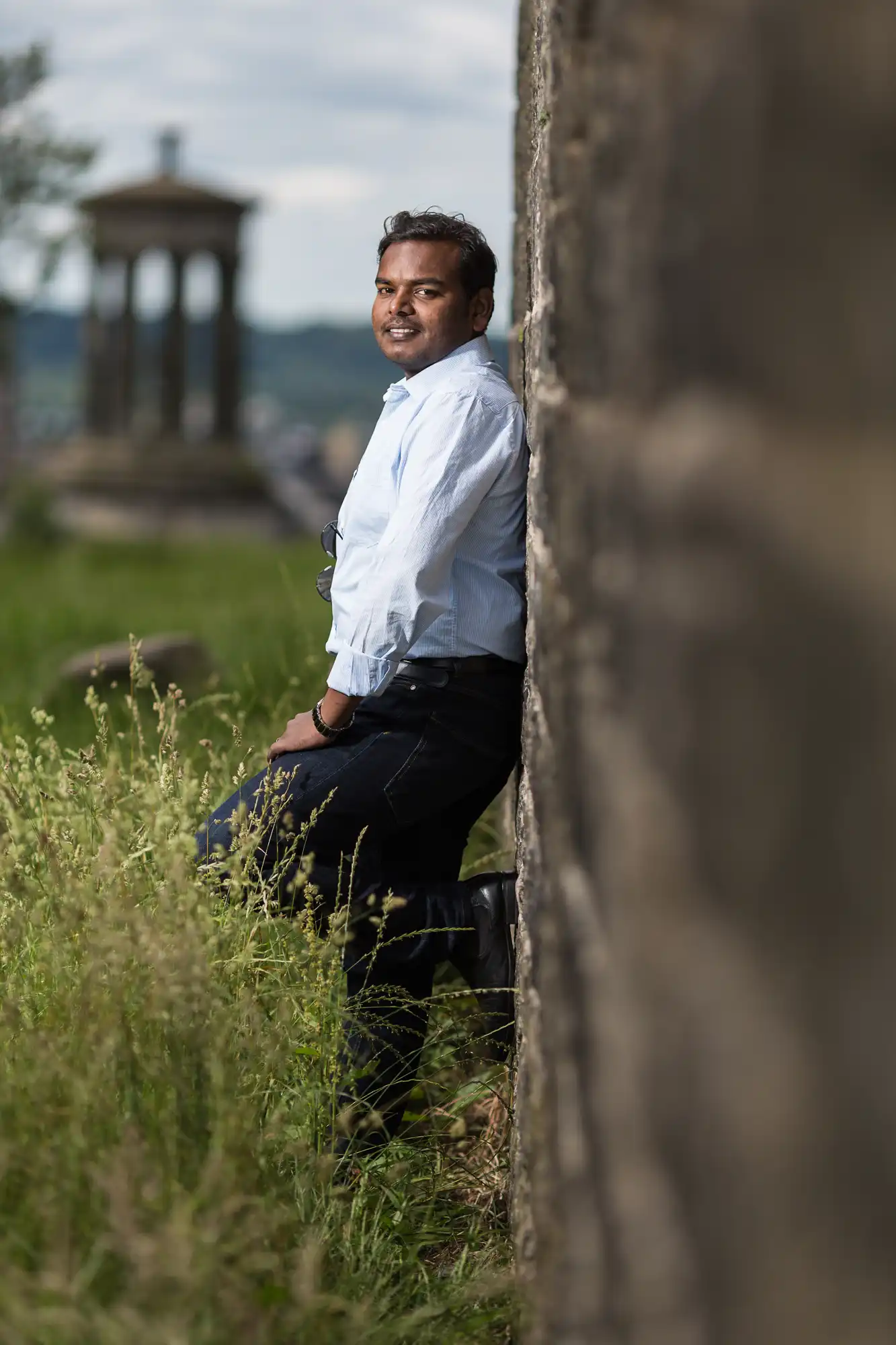 A person in a light blue shirt and dark pants leans against a stone wall in an outdoor area with tall grass and a structure in the background.