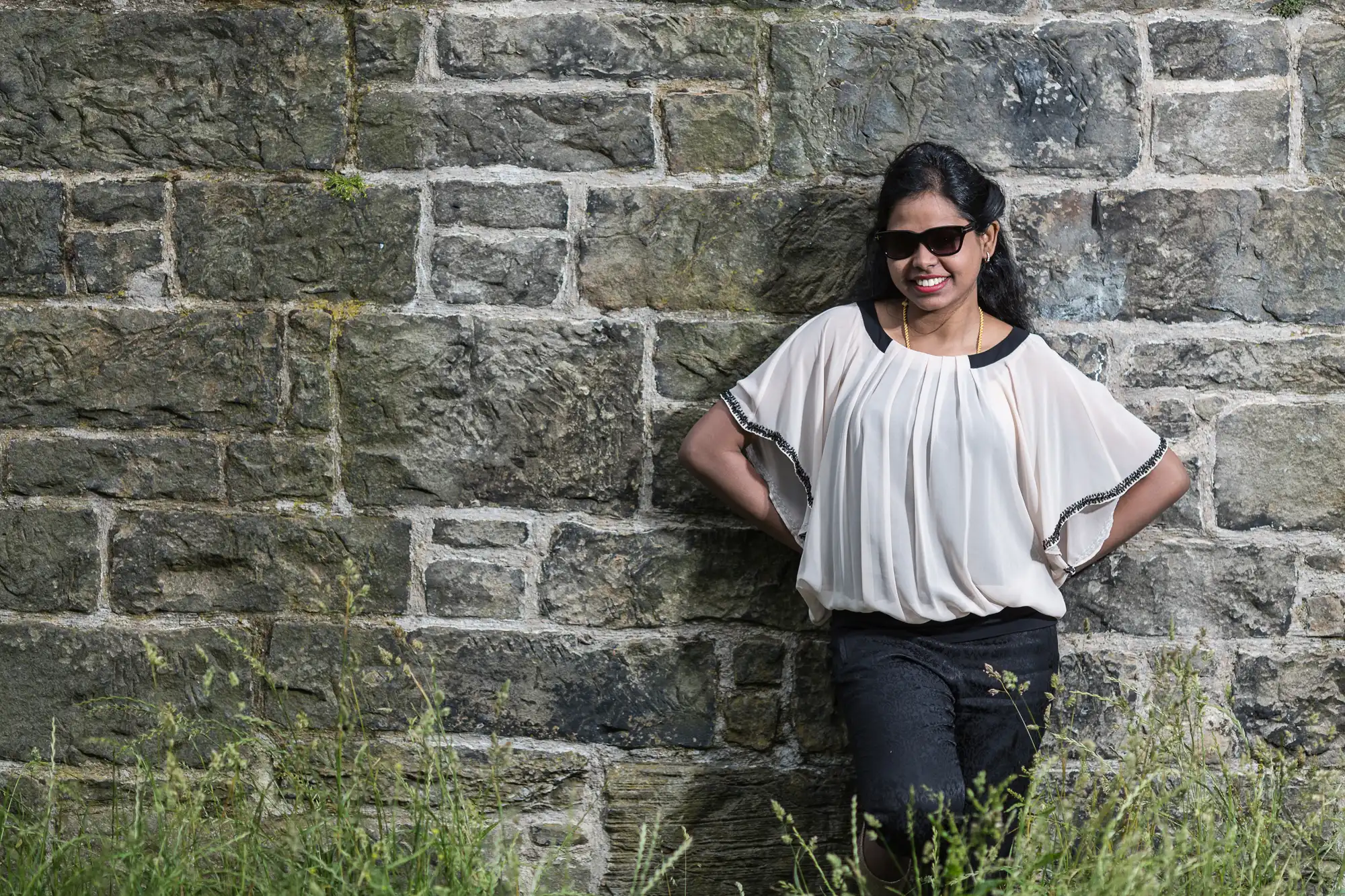 A woman in a white blouse and black pants stands against a stone wall with a slight smile. She is wearing sunglasses and has her hands on her hips. There is grass in the foreground.