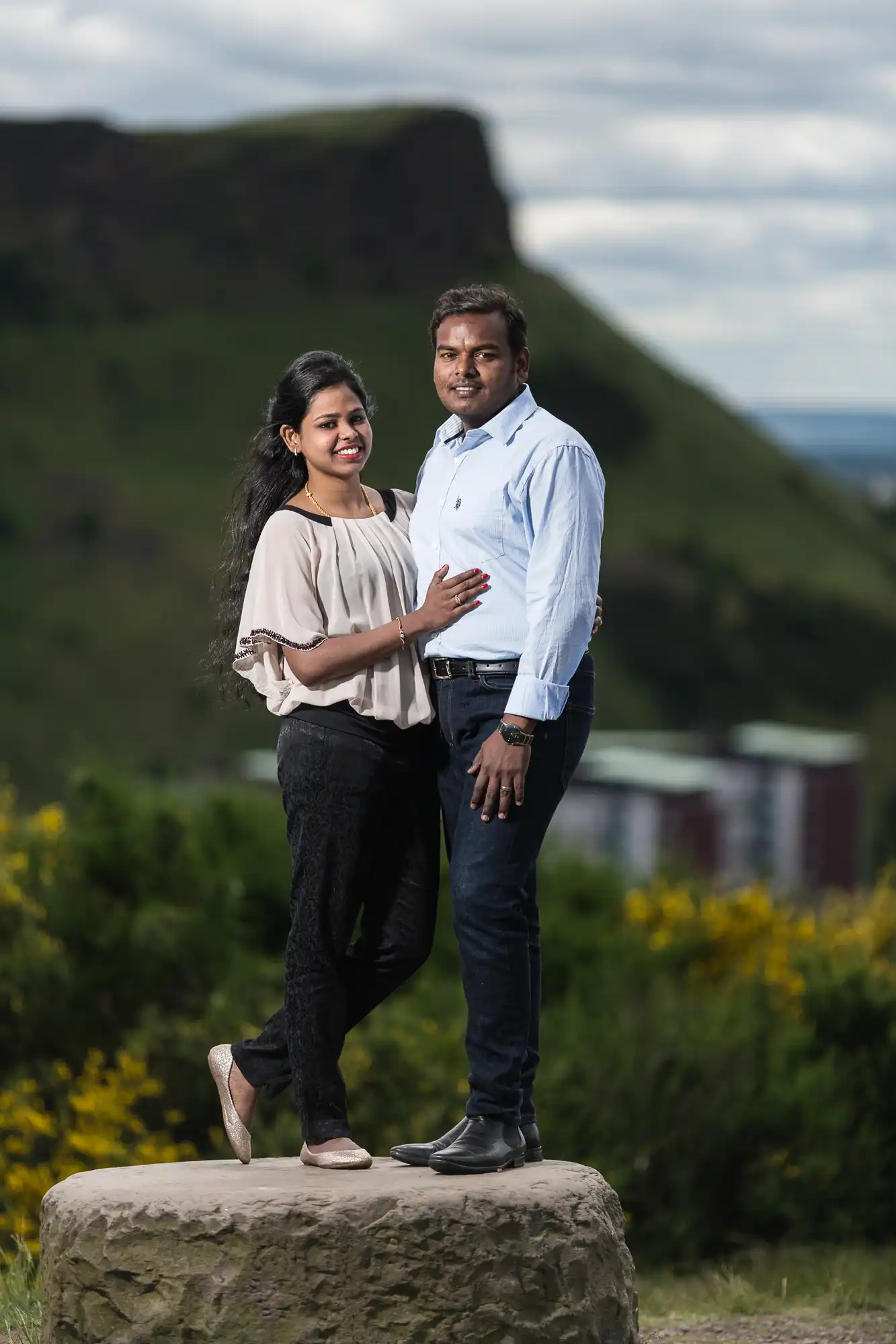 A couple stands on a stone platform with a hilly landscape in the background, posing closely together while smiling at the camera.